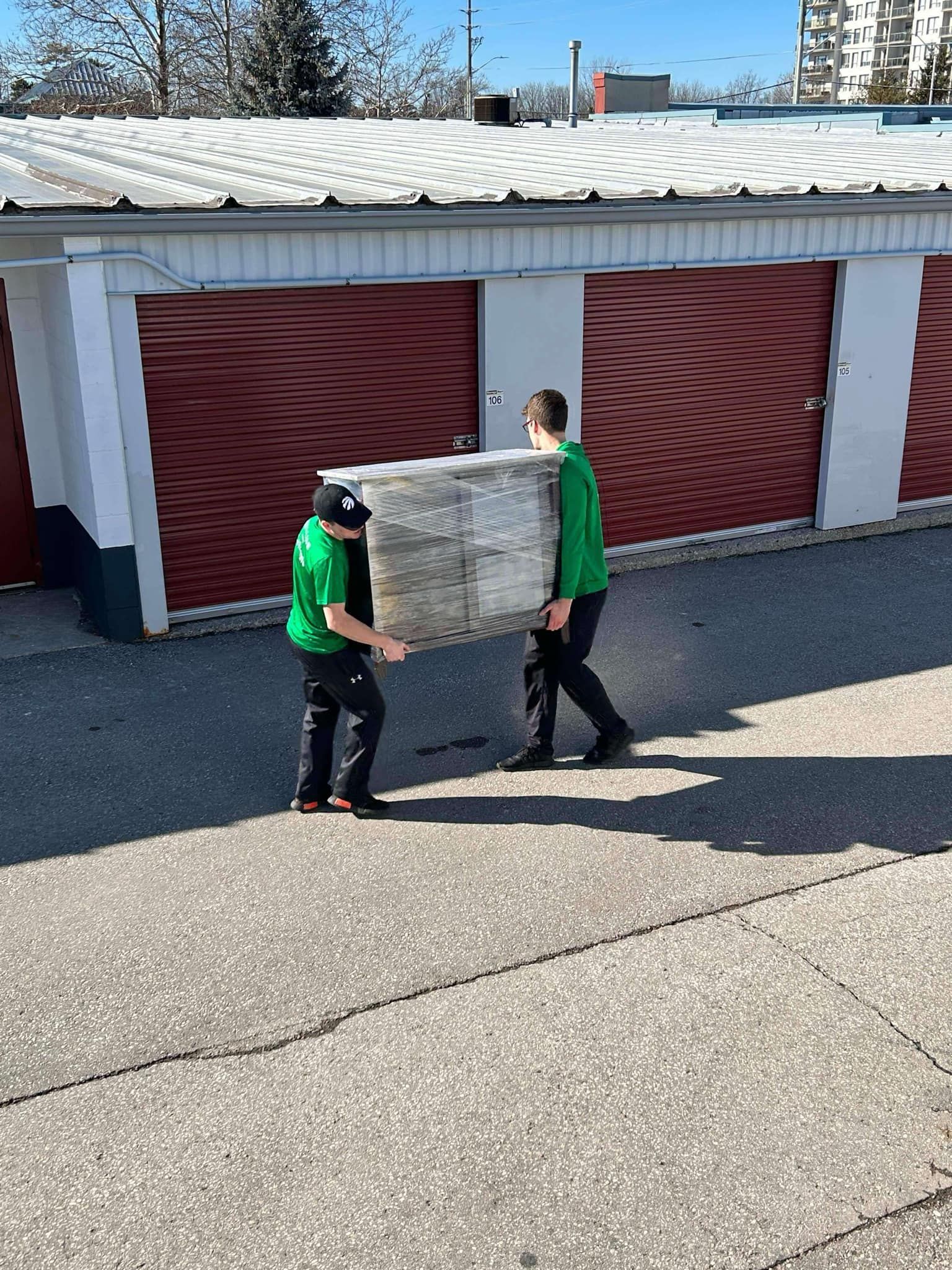 Two people in green shirts carrying a large, light-colored rectangular item in front of storage units.