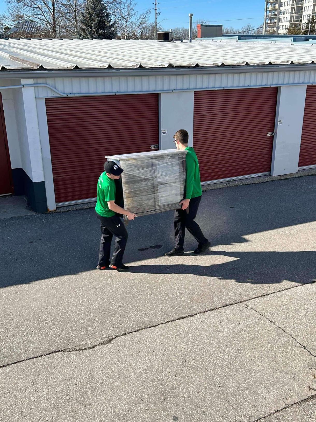 Two workers in green shirts carry a large bundled item across a storage facility parking lot.