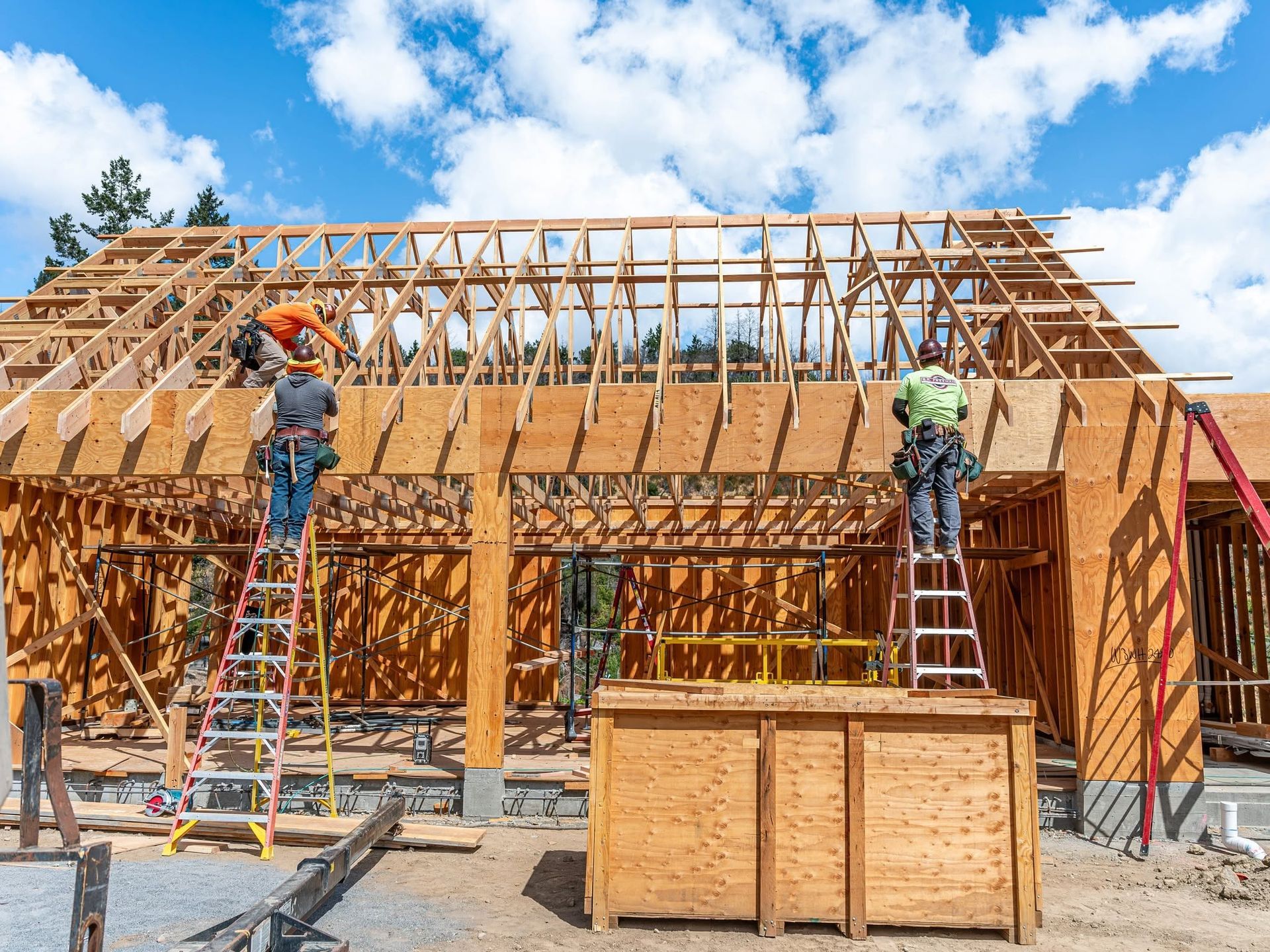 Workers building a wooden roof frame on a house under construction, with ladders and a blue sky.