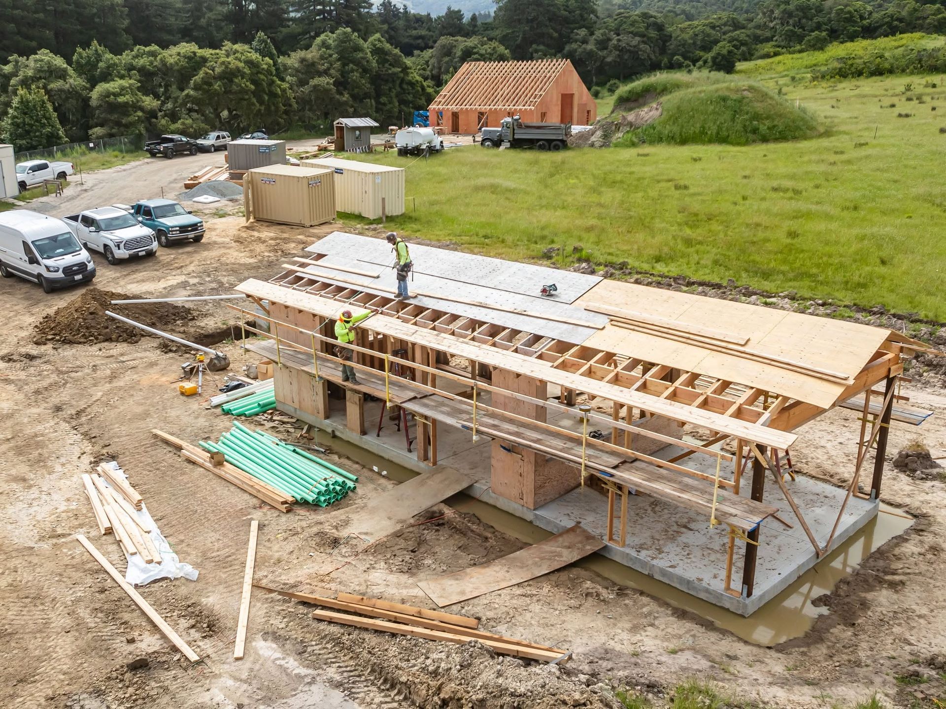 Construction site with partially built wooden house and workers on the roof, beside parked cars and grassy field