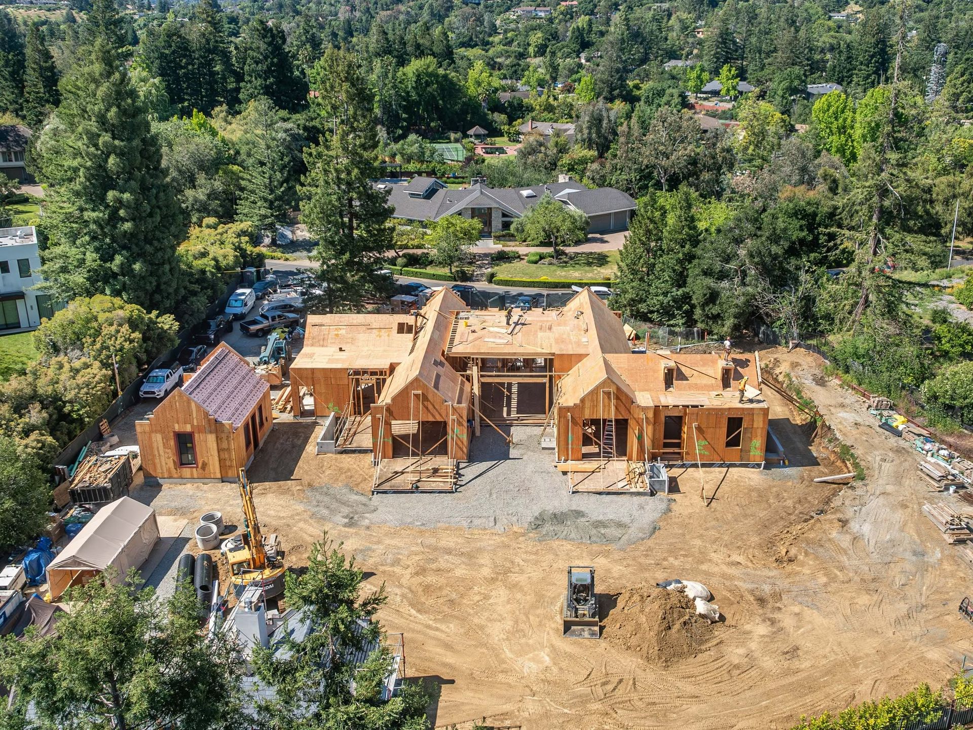 Aerial view of a house under construction with exposed wooden framing on a cleared lot surrounded by trees.