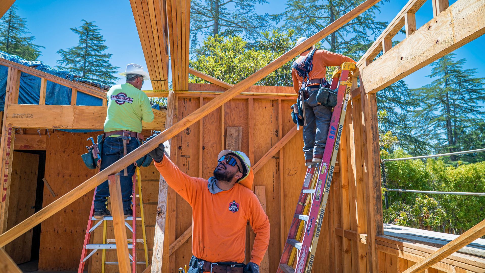 Construction workers framing a wooden house, one on a ladder and others lifting beams under a blue sky