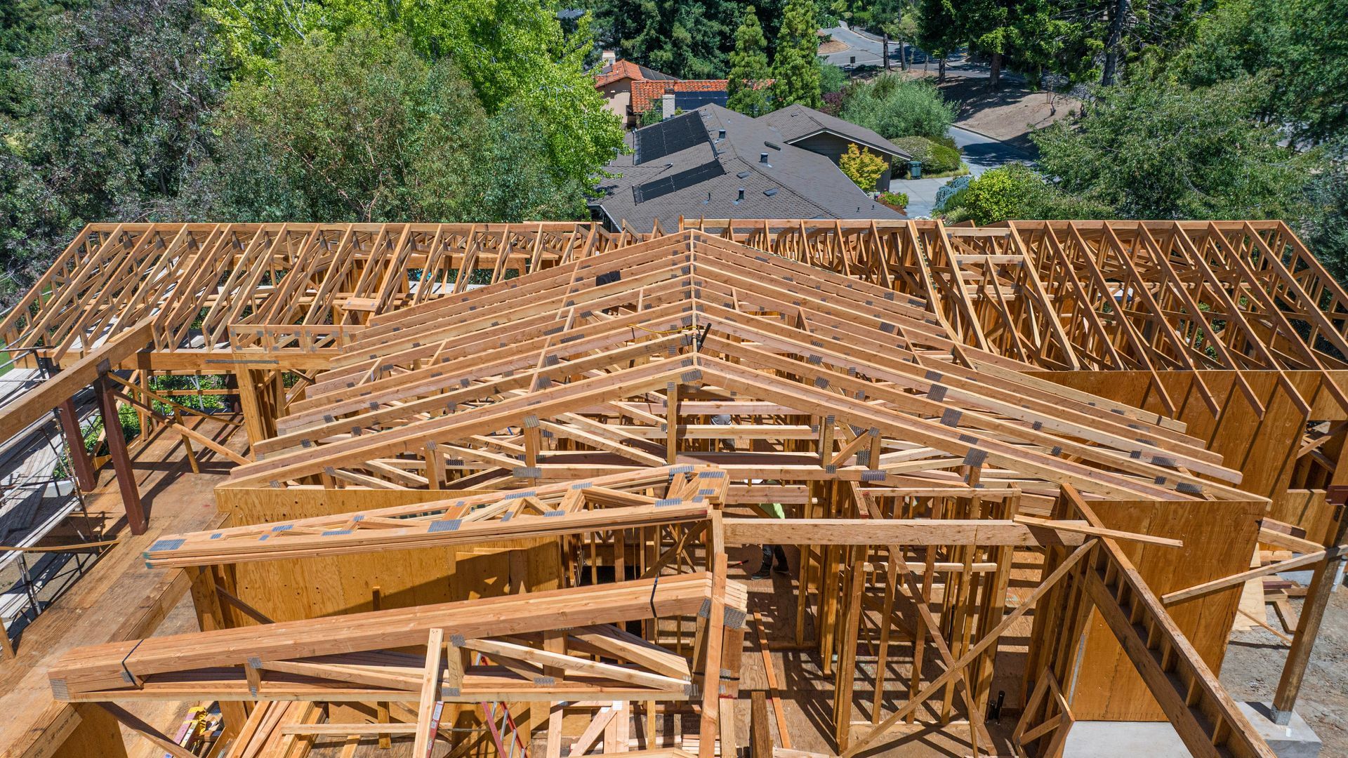 Wooden house roof frame under construction with trees and neighboring homes in the background