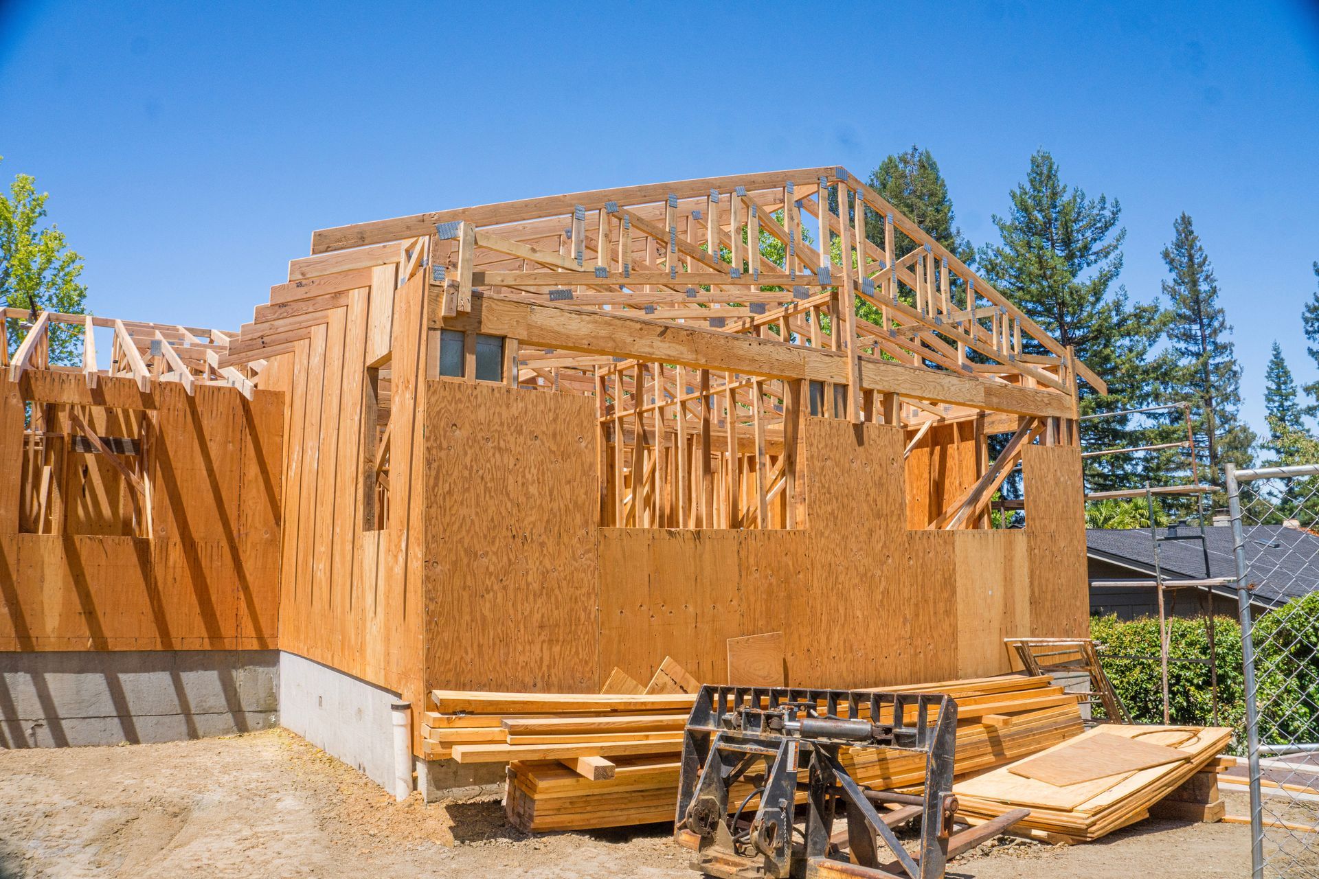 Wood-framed house under construction on a sunny site with lumber stacks and blue sky