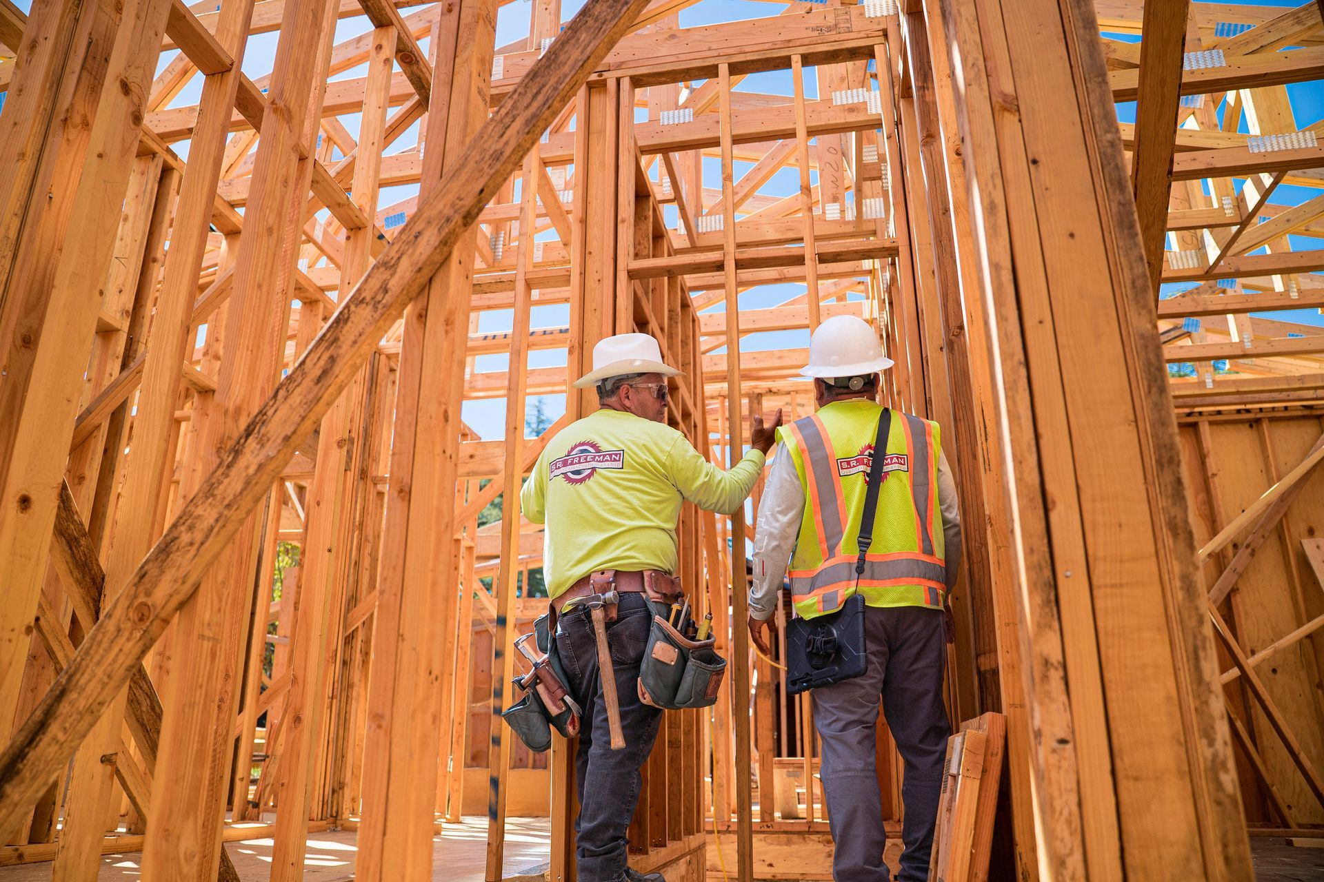 Two construction workers in safety vests and hard hats frame a wooden house under construction.