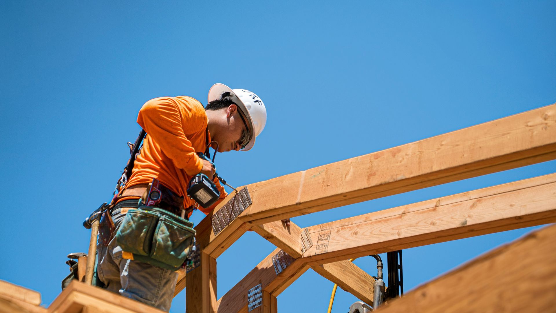 Construction worker in orange safety gear fastening wooden roof beams against a blue sky