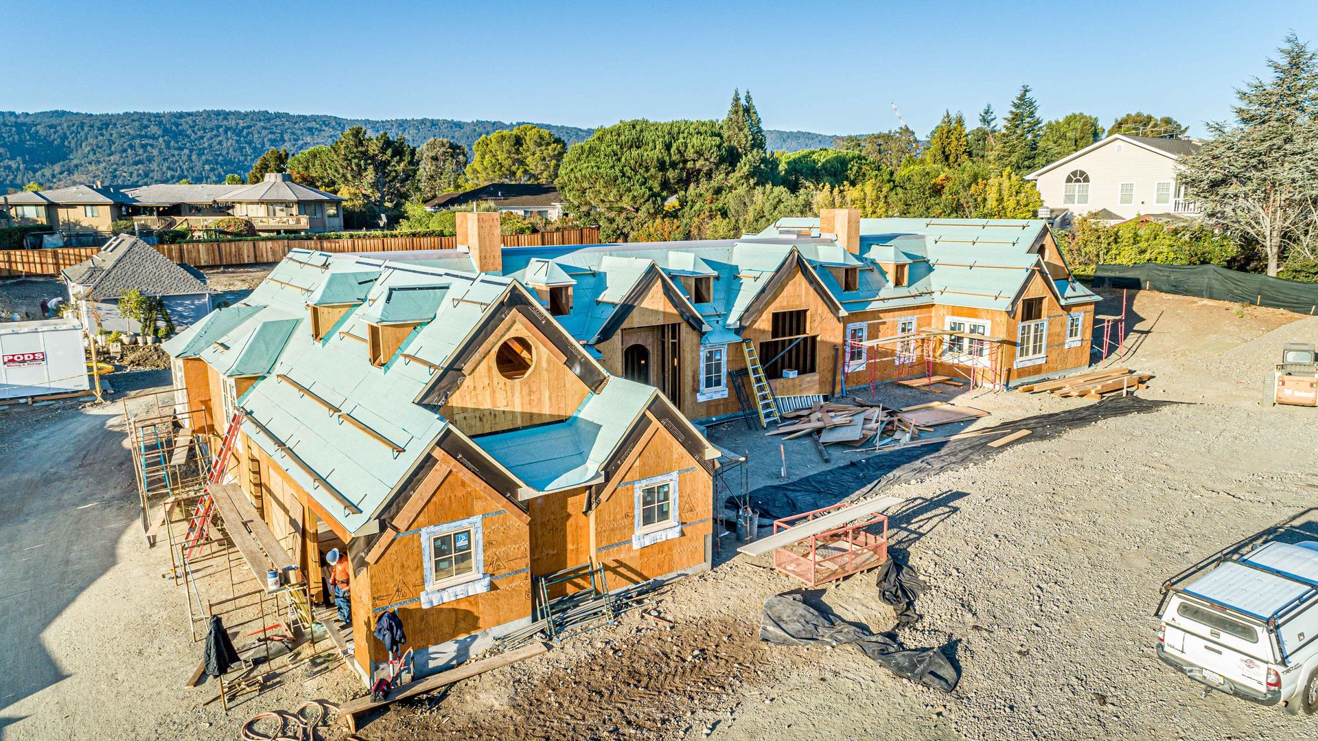 New houses under construction with blue roofs and wood siding in a gravel lot, viewed from above