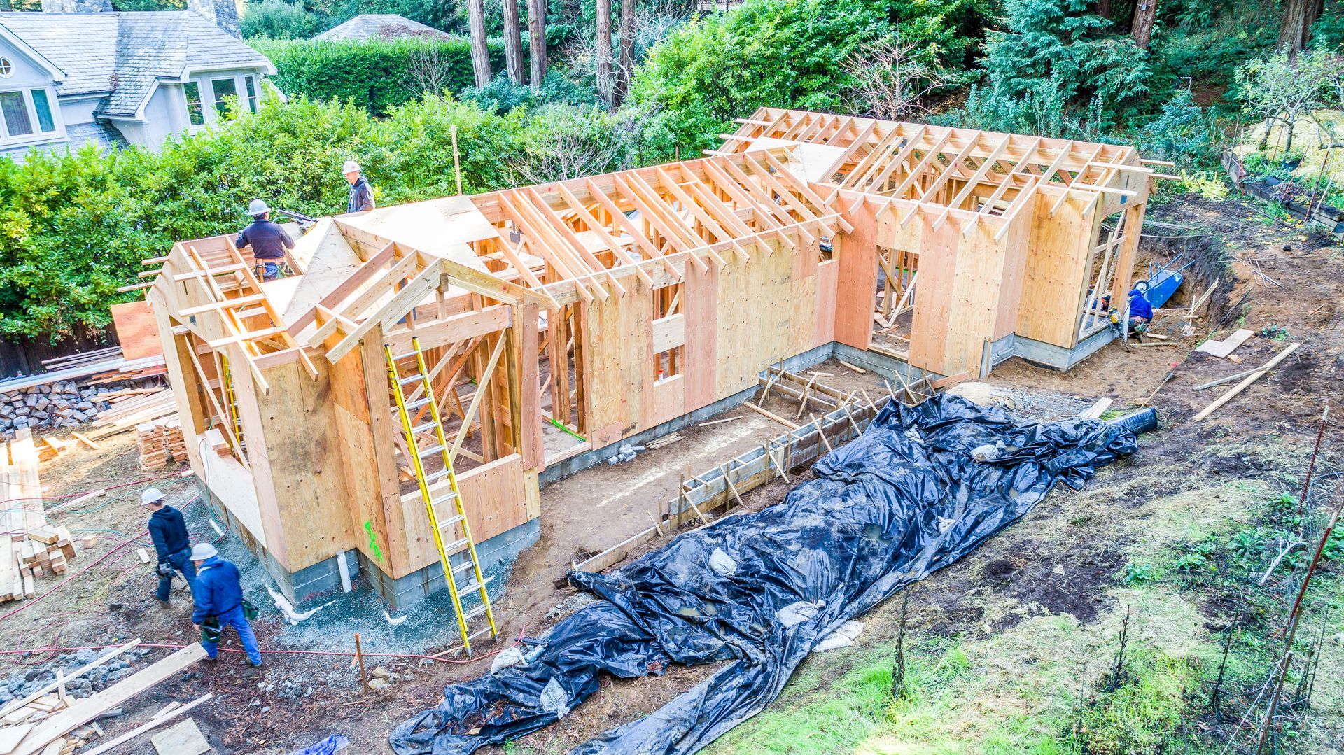 Wood-frame house under construction with workers and ladder on a dirt lot surrounded by trees
