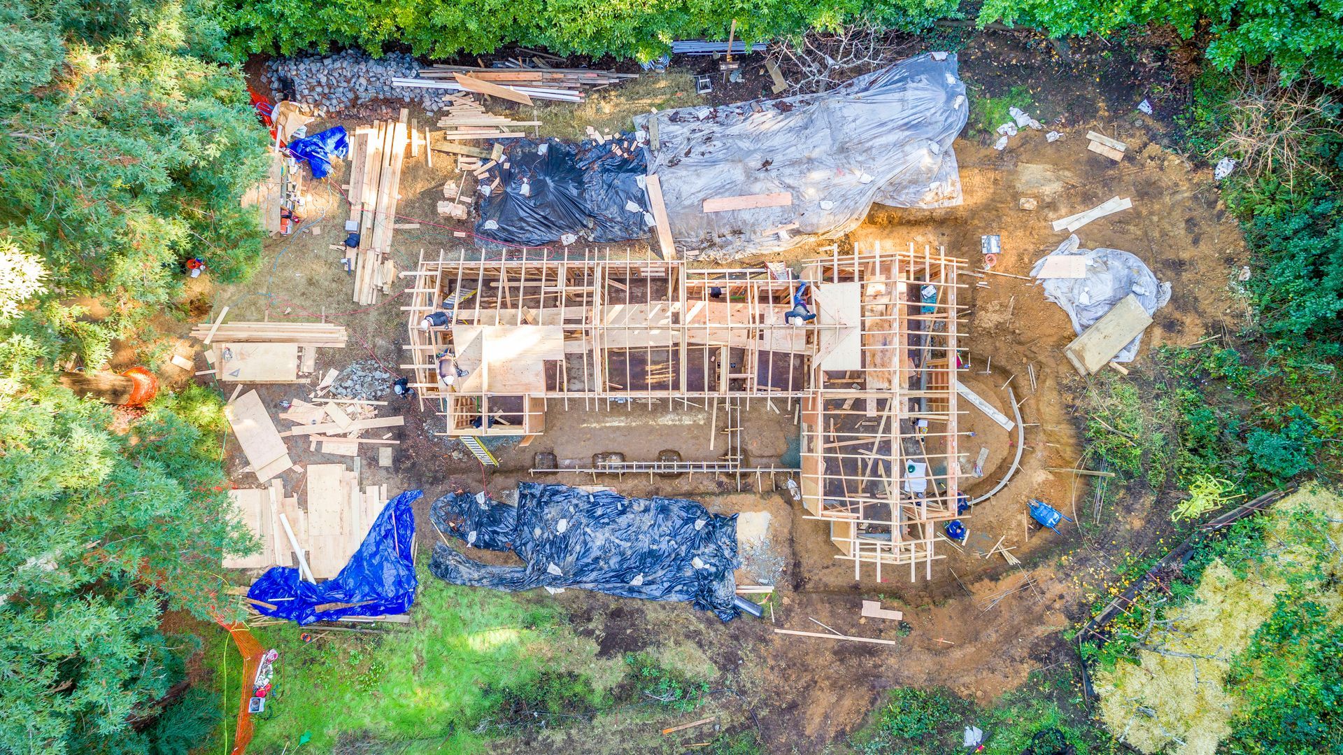 Aerial view of a house under construction surrounded by trees, with roof framing and blue tarps.