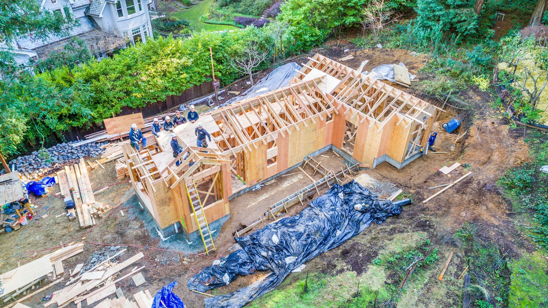 Wooden house under construction on a sloped, wooded hillside, with blue tarp and exposed framing.