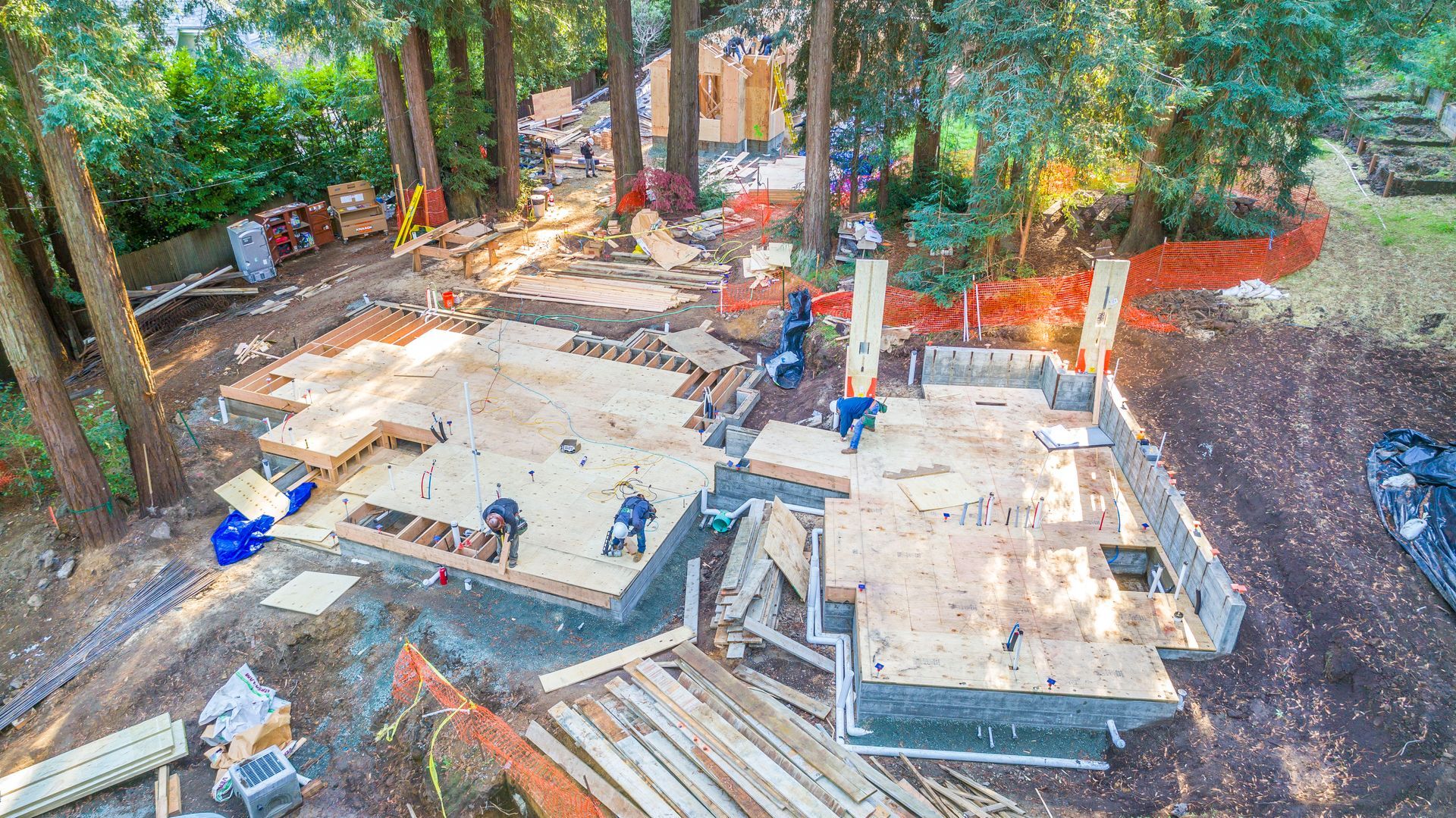 Construction site with concrete foundation, workers, and stacked lumber amid trees and dirt