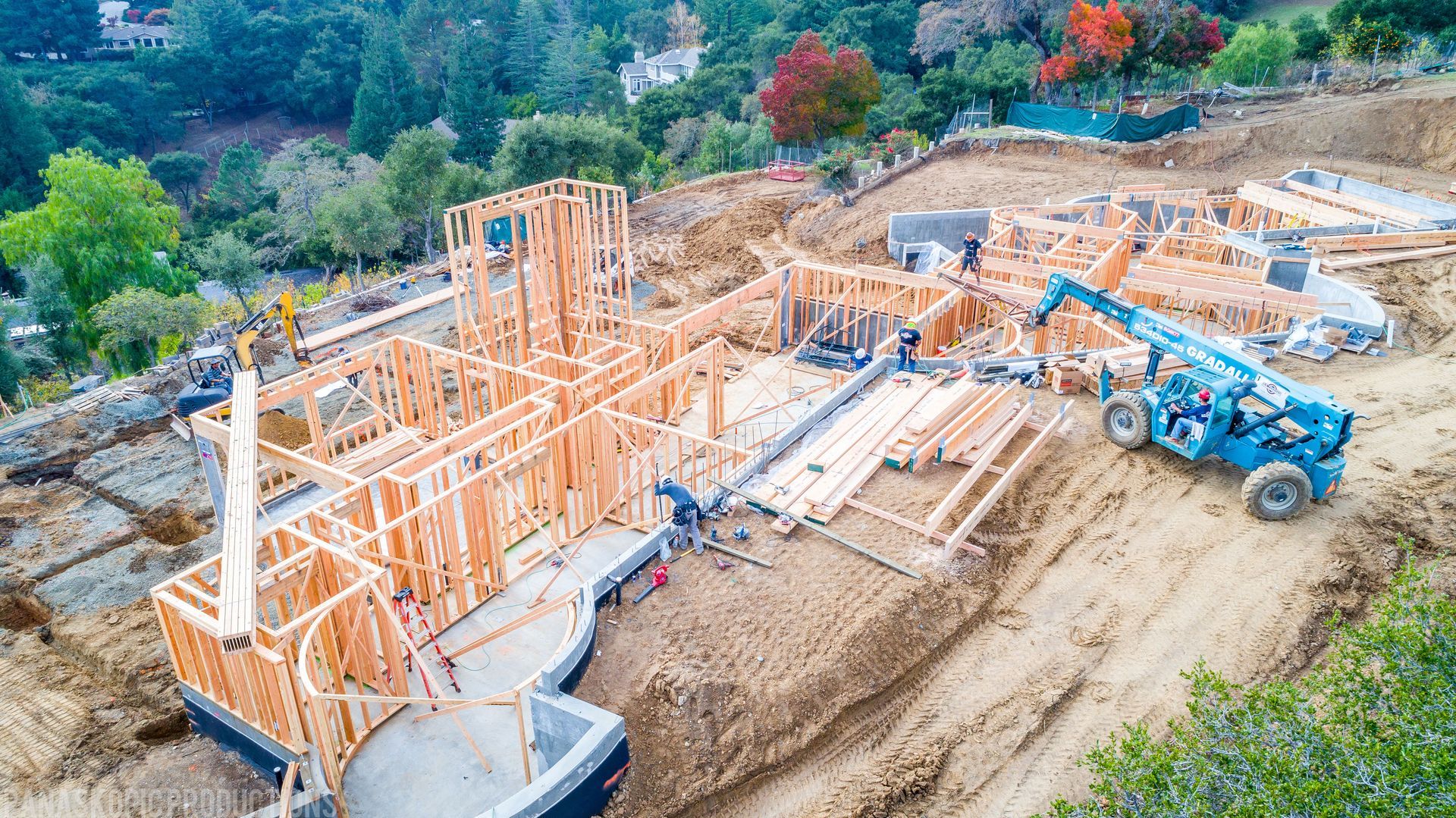Partially built wooden house framing on a hillside with blue construction equipment and dirt roads