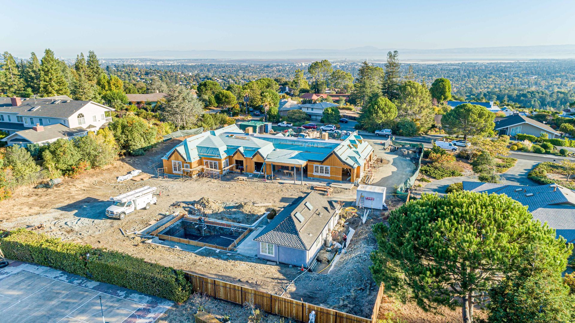 Aerial view of a hillside home construction site with blue roofs, excavated foundation, and city skyline in the distance
