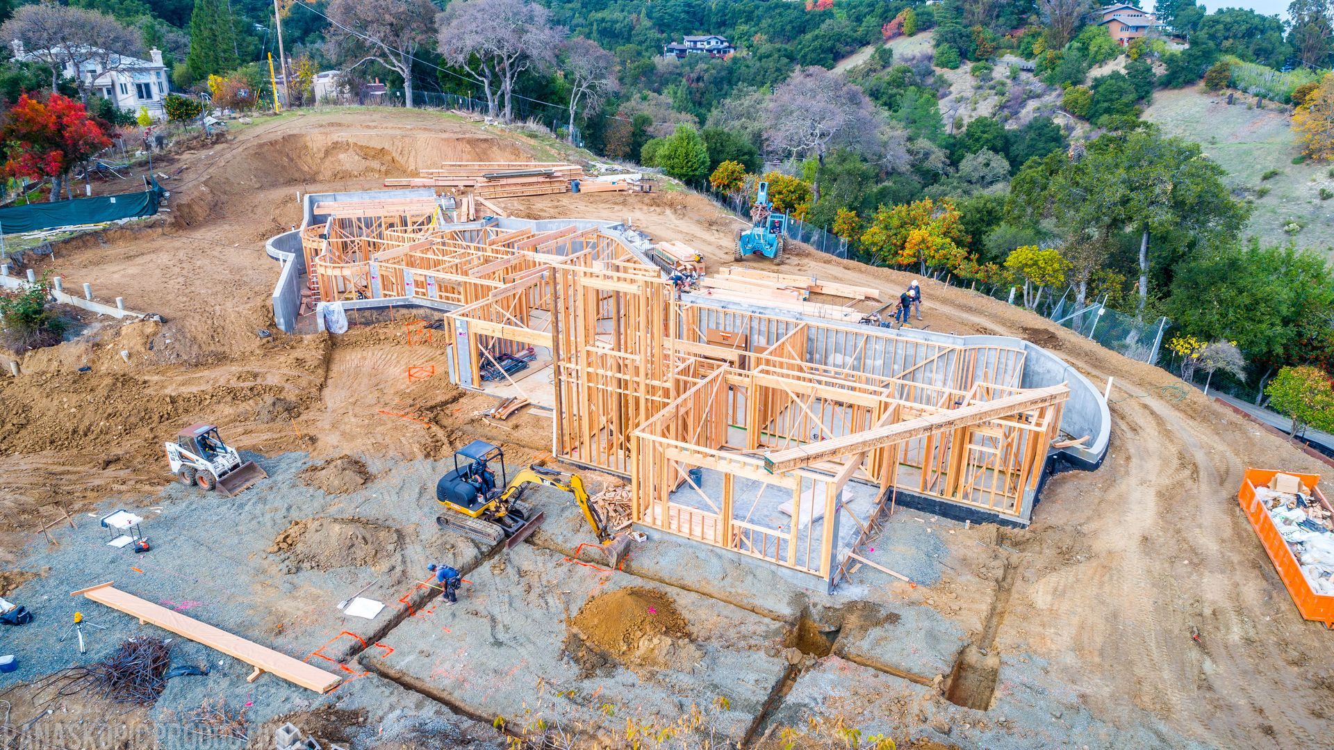 Wood-framed house under construction on a cleared hillside, surrounded by trees and dirt.