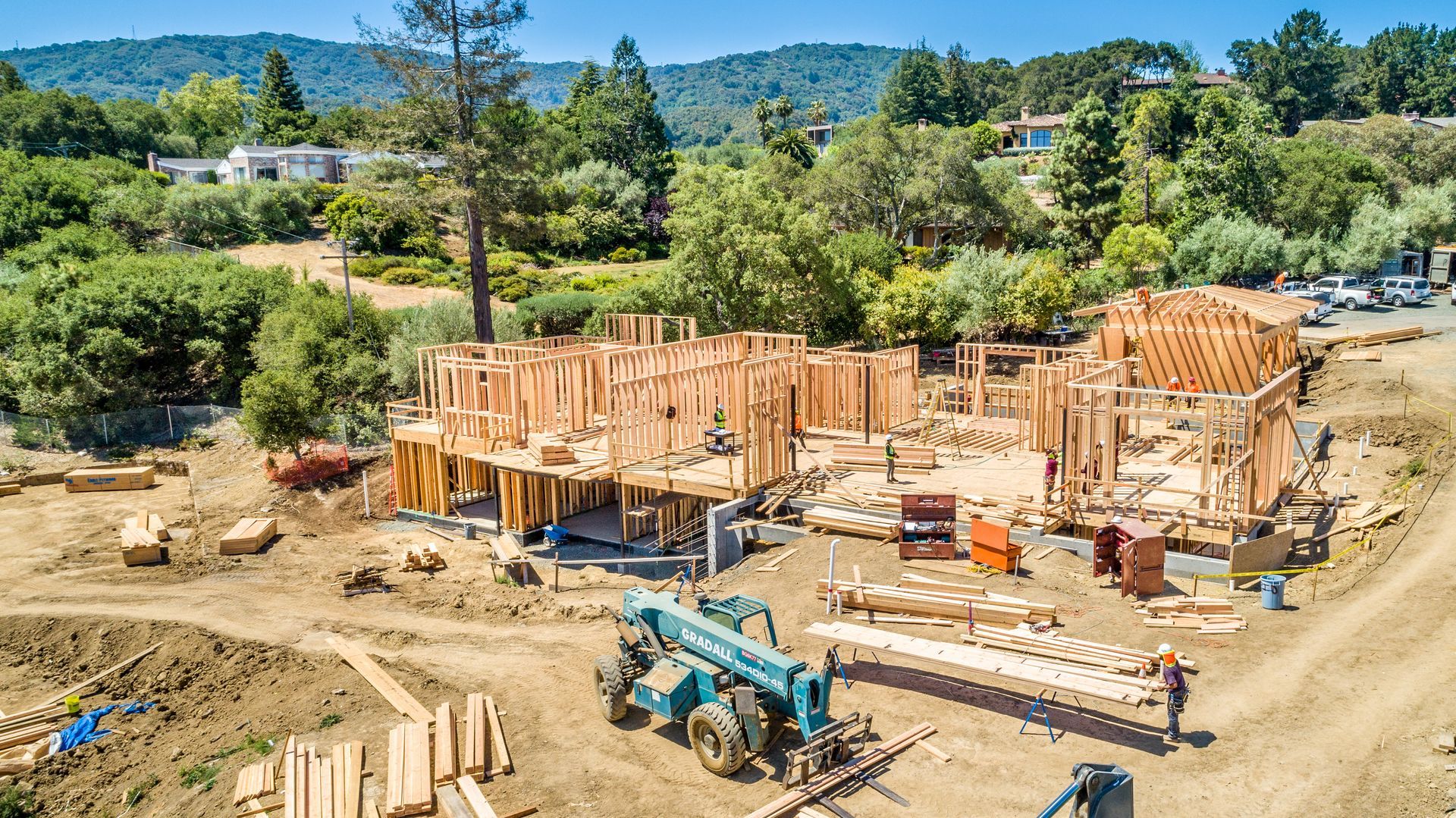 Construction site with wooden house framing, excavator, and workers on a cleared lot under a sunny sky