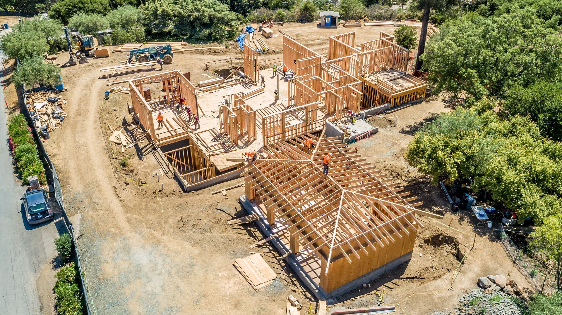 Aerial view of a house under construction with wooden framing and dirt access roads surrounded by trees