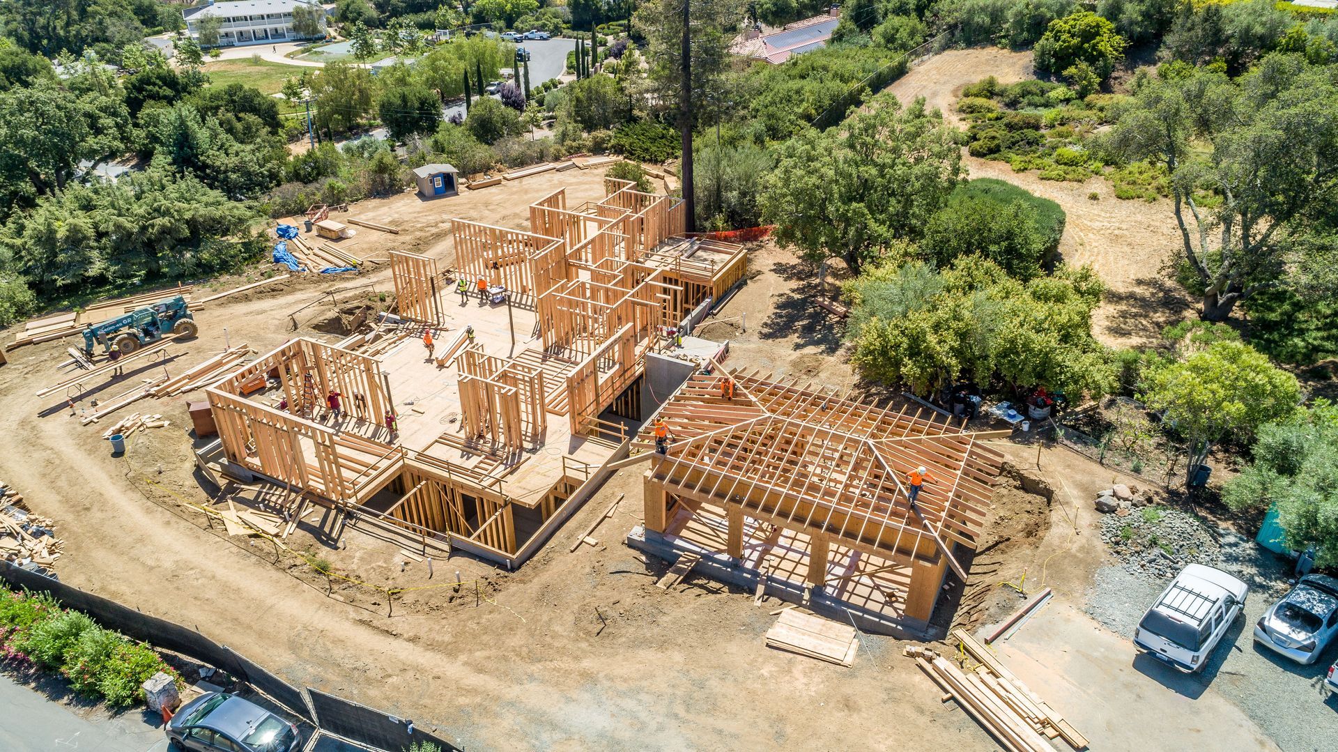 Aerial view of a house under construction with wooden framing, surrounded by trees and dirt roads.