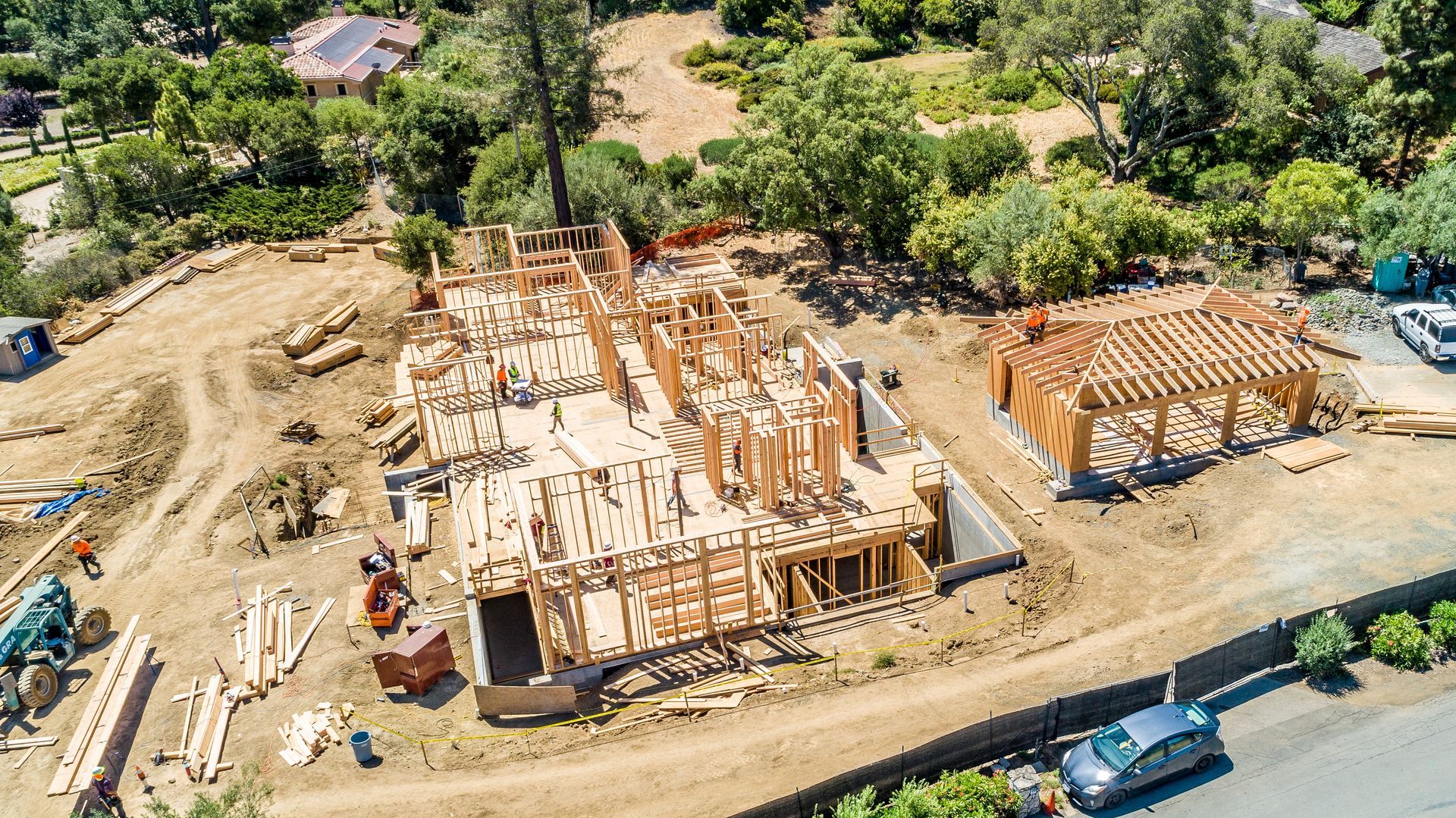 Aerial view of a house under construction with wooden framing on a cleared site beside a road and trees