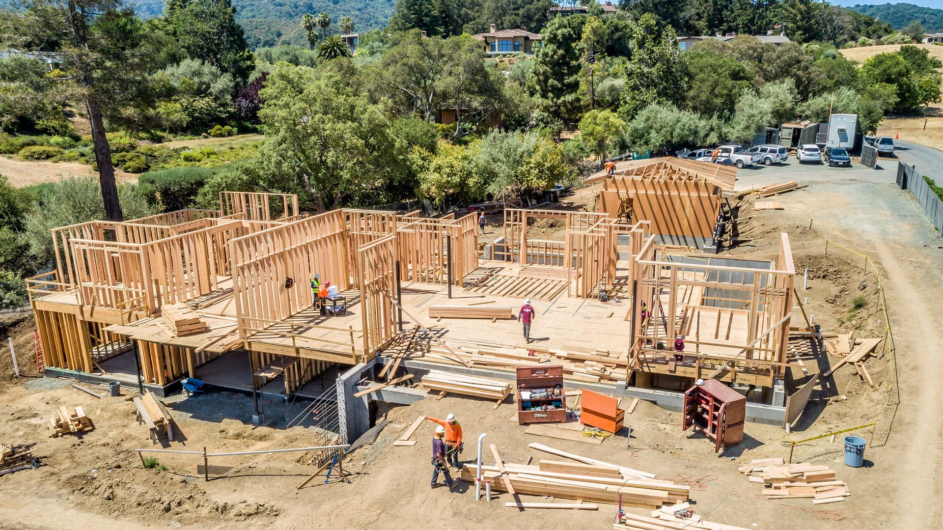 Wood-framed house under construction on a dirt lot with workers, lumber, and heavy equipment around it