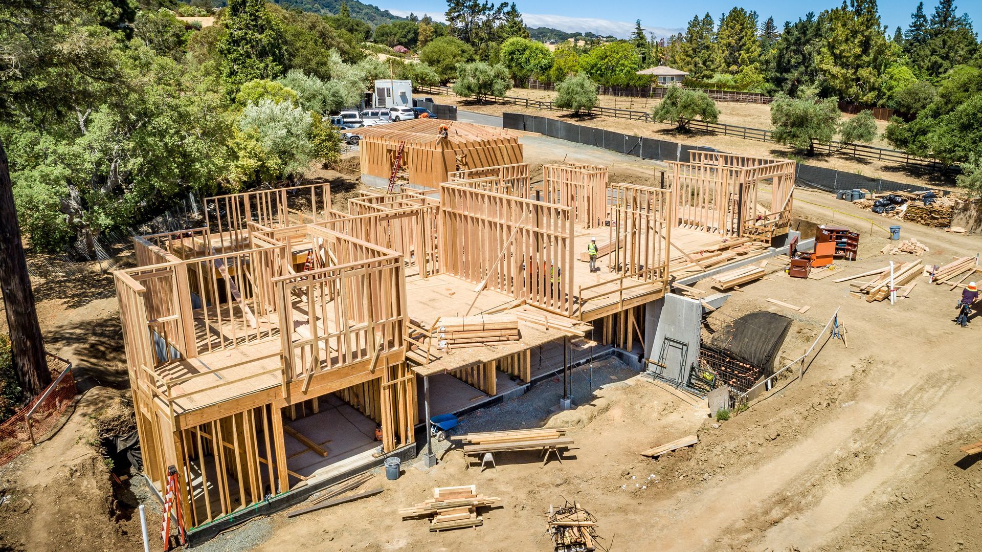 Wood-framed house under construction on a dirt lot, with scaffolding and a wooded hillside in the background