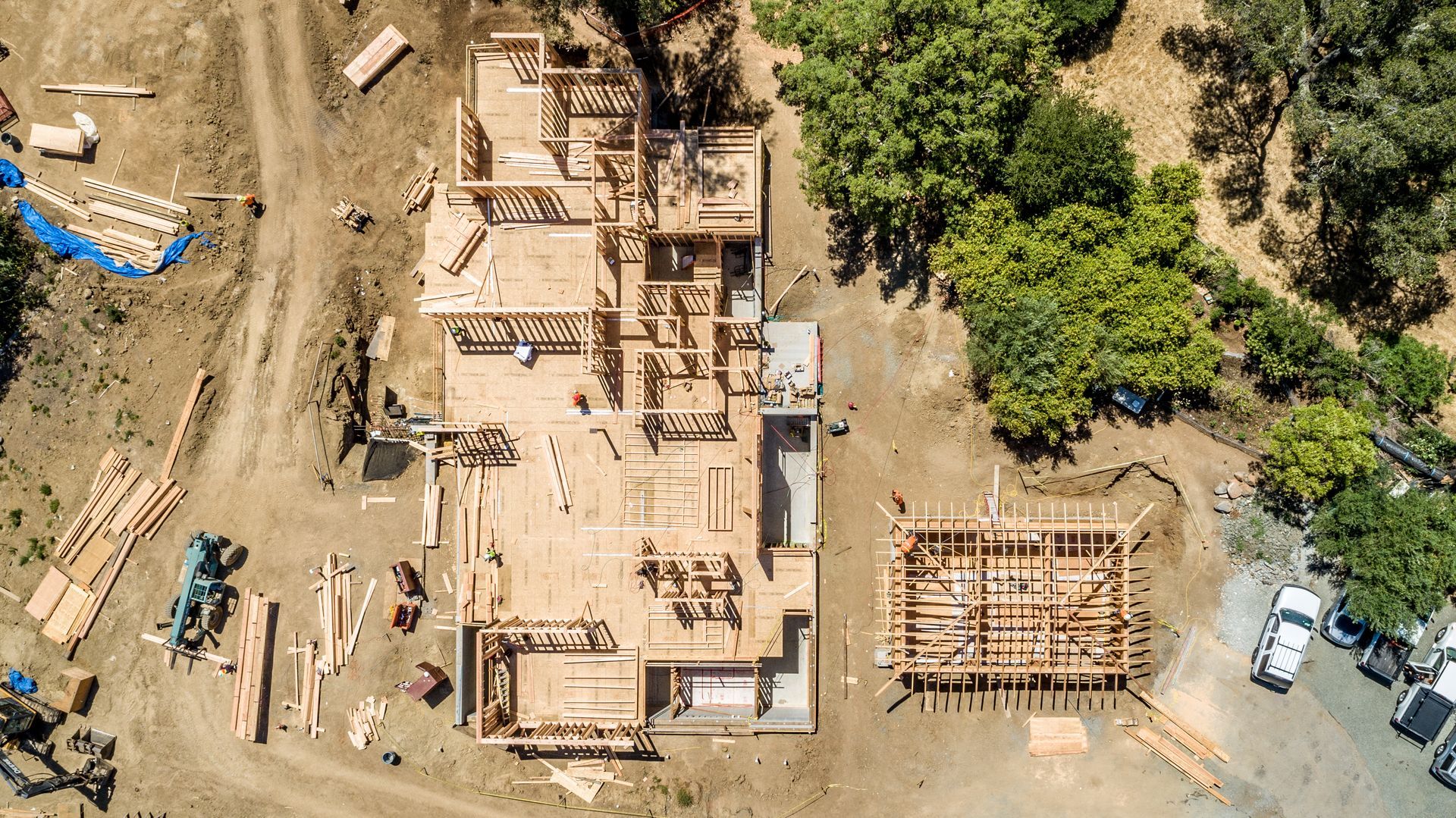 Aerial view of a construction site with building foundations, heavy equipment, and surrounding trees and dirt roads