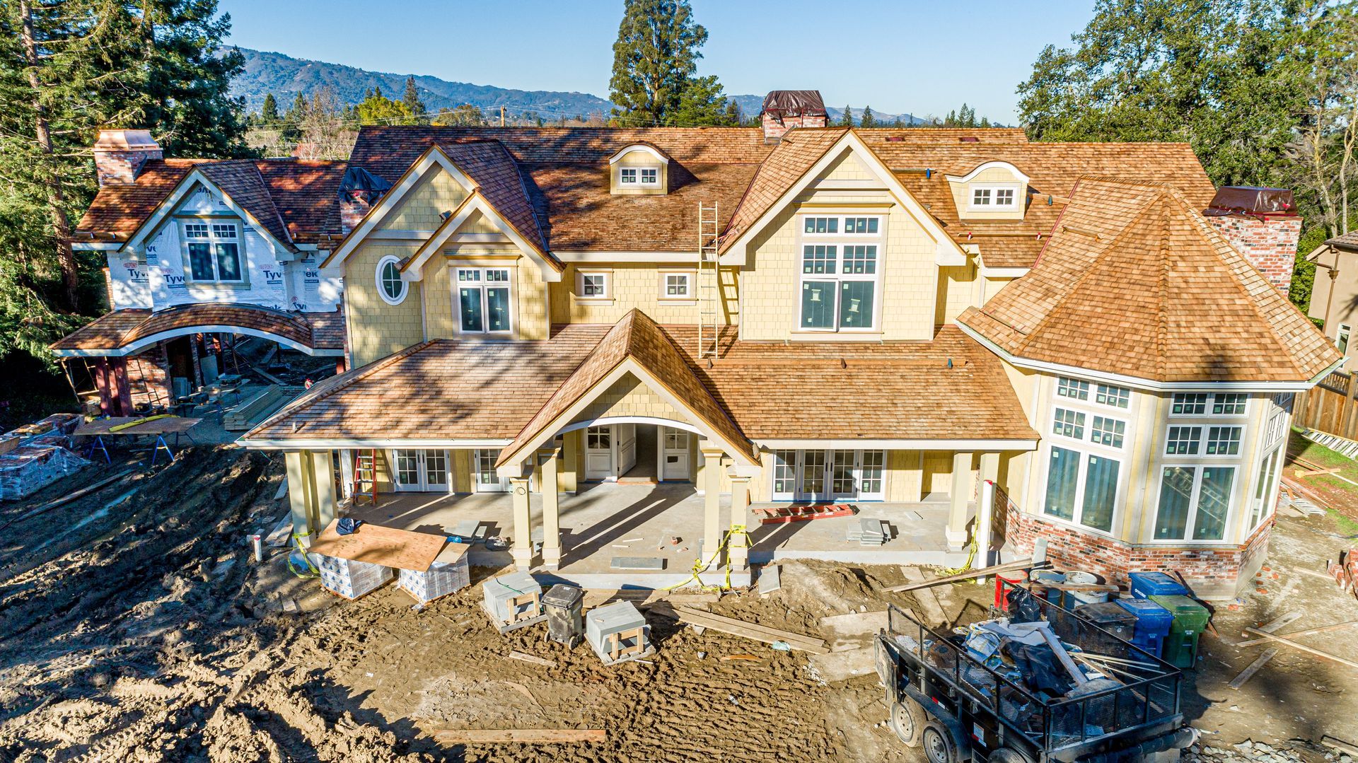 Large house under construction with tan siding, dormer windows, and exposed dirt in front yard