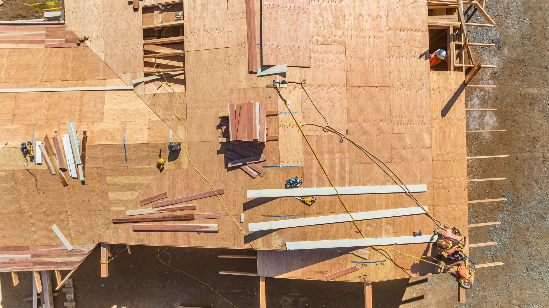 Overhead view of a plywood-framed roof under construction with workers and stacked lumber.