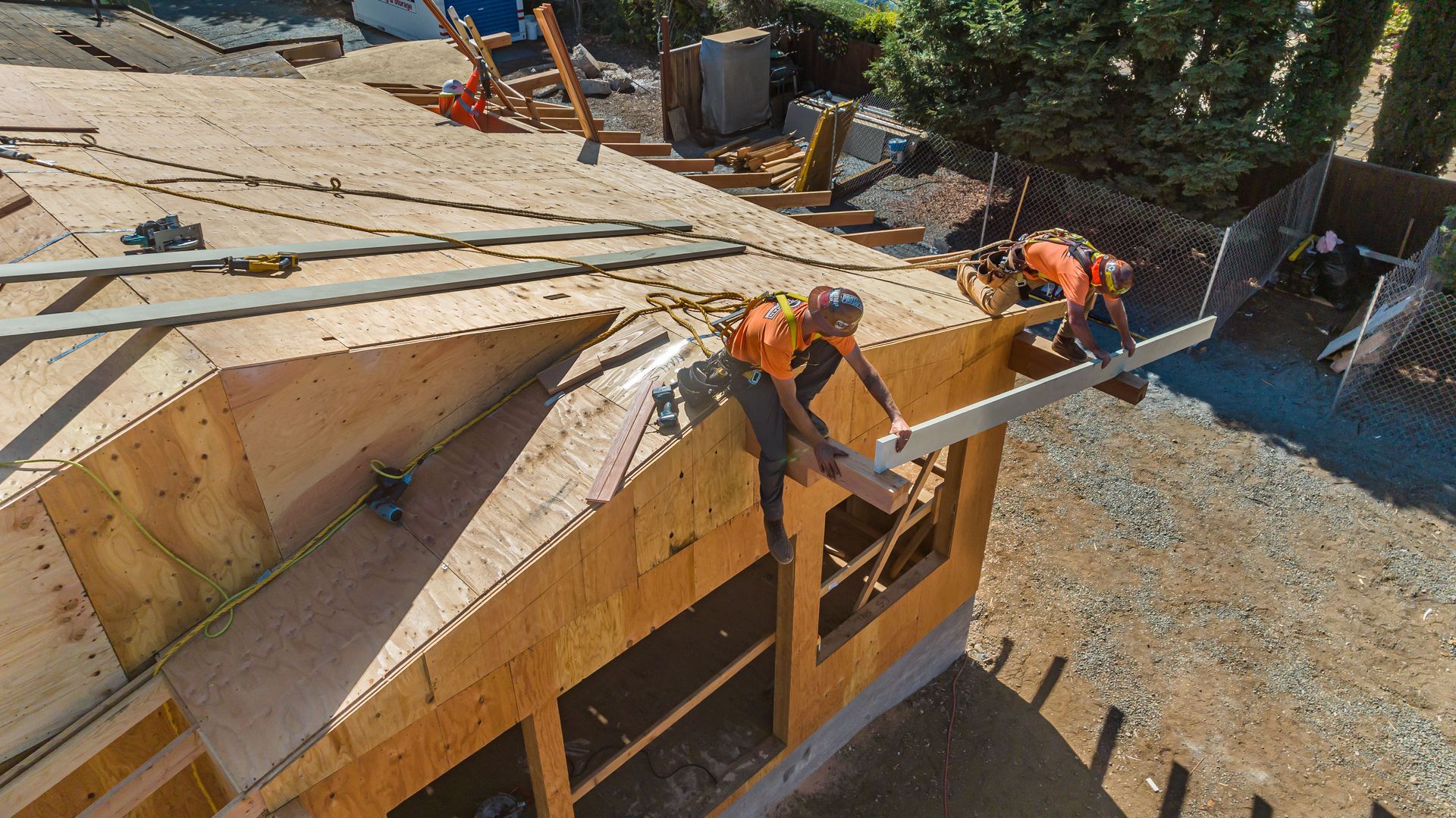 Workers on a wooden house roof installing framing, with lumber stacked around the construction site.
