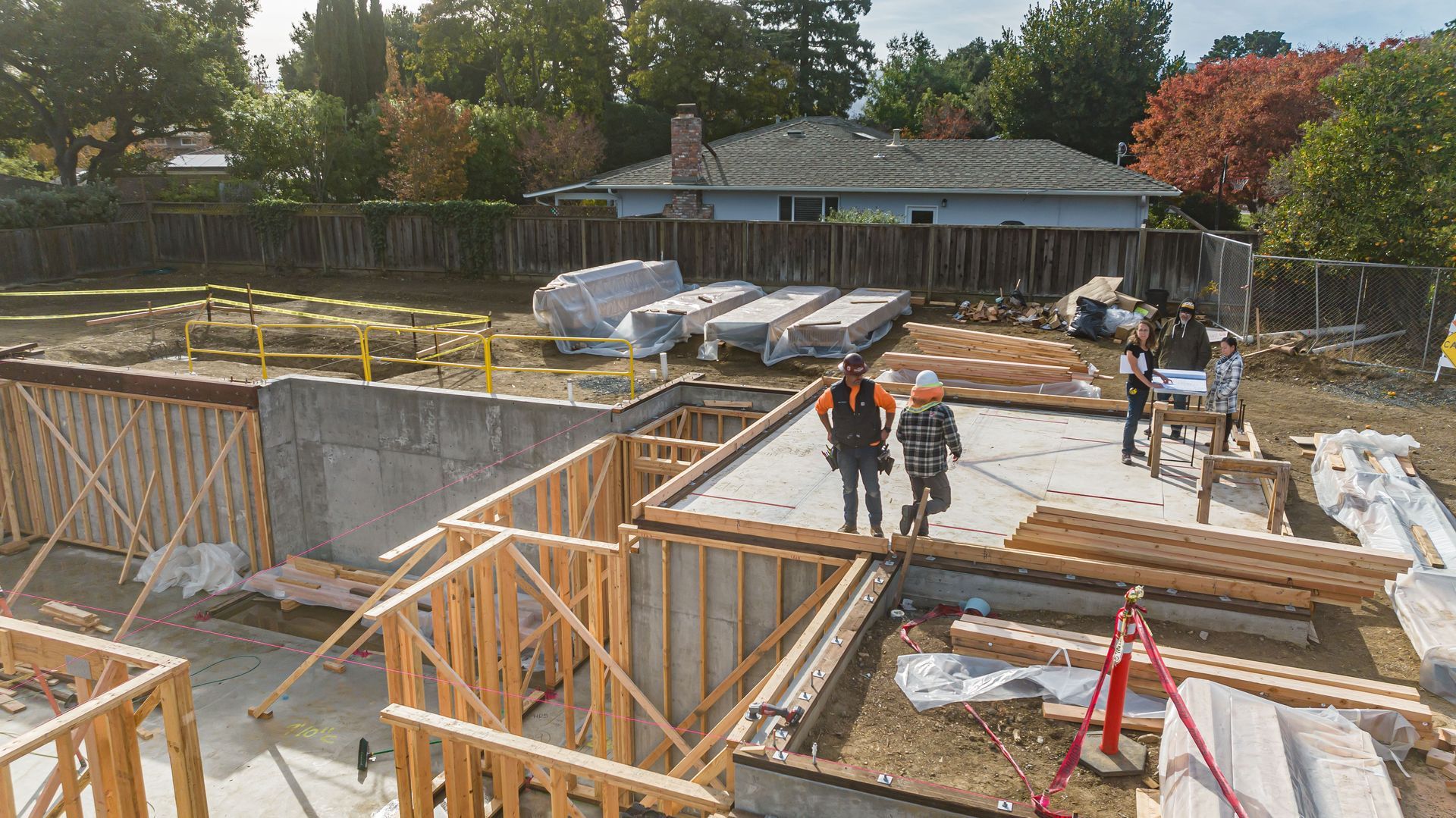 Construction site with workers, concrete foundation walls, wooden framing, and vehicles in the background