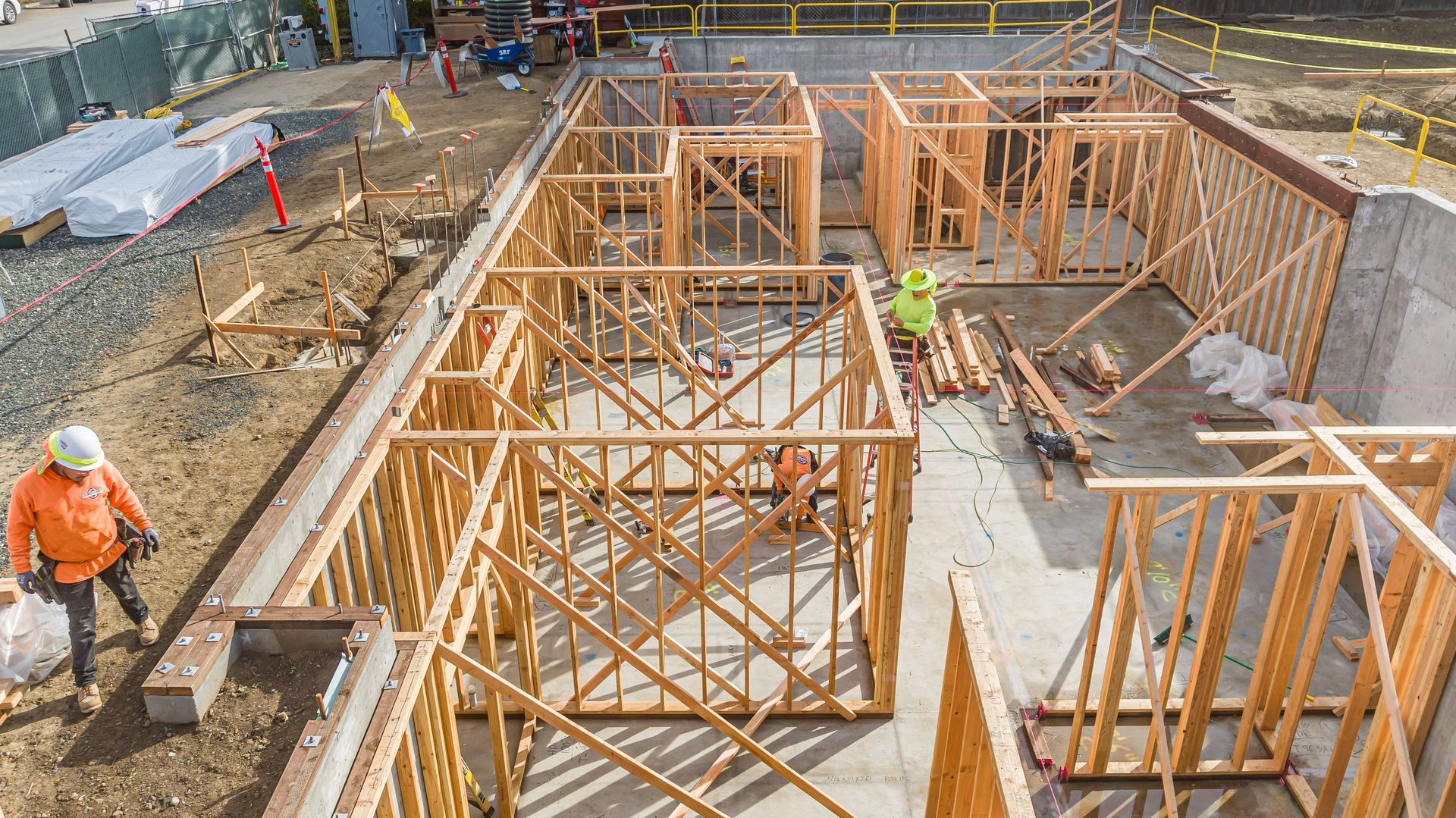 Construction site with wooden foundation forms and scaffolding, worker in orange vest at left