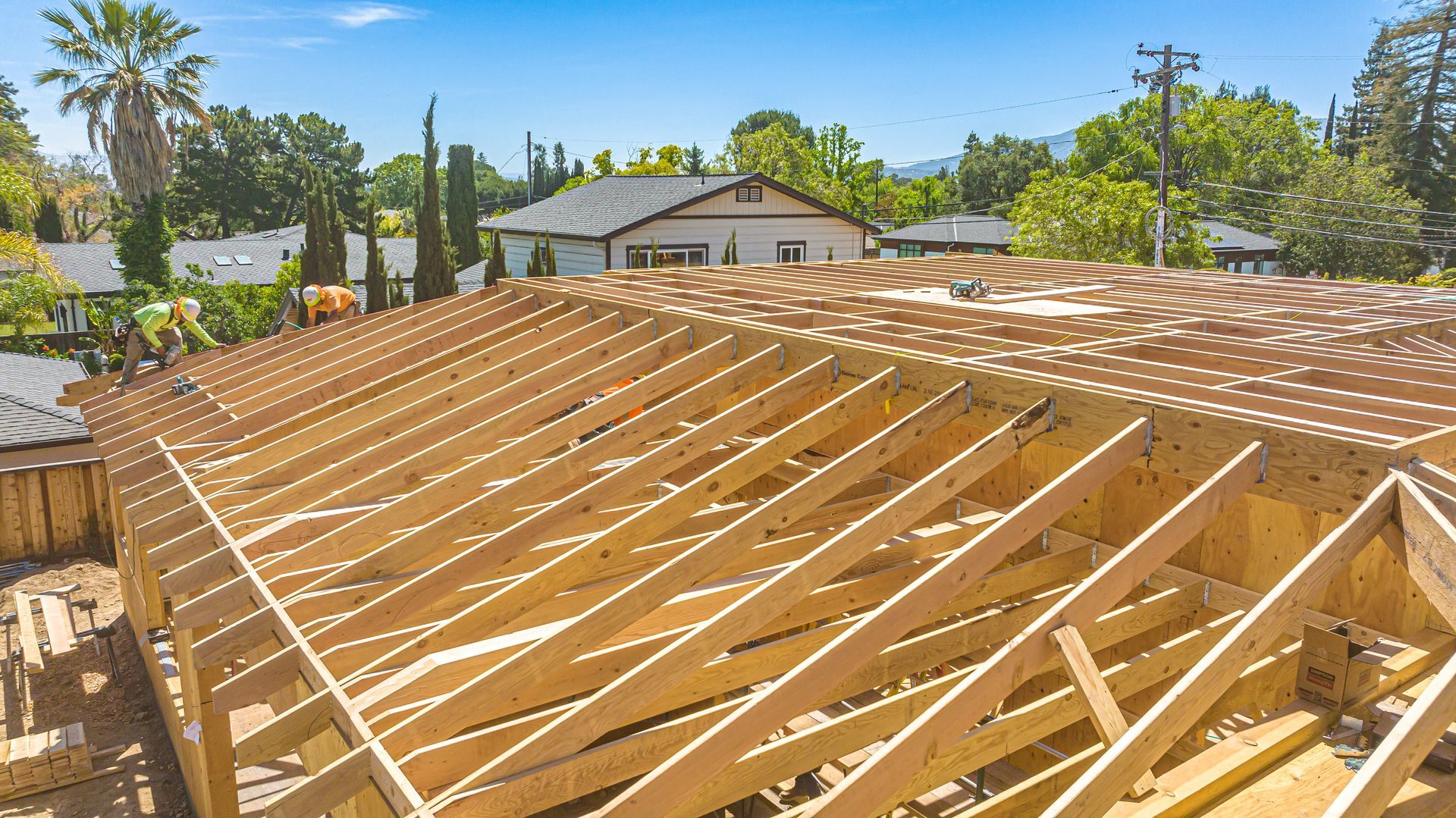 Wooden roof framing under construction on a sunny residential site, with houses and palm trees in the background