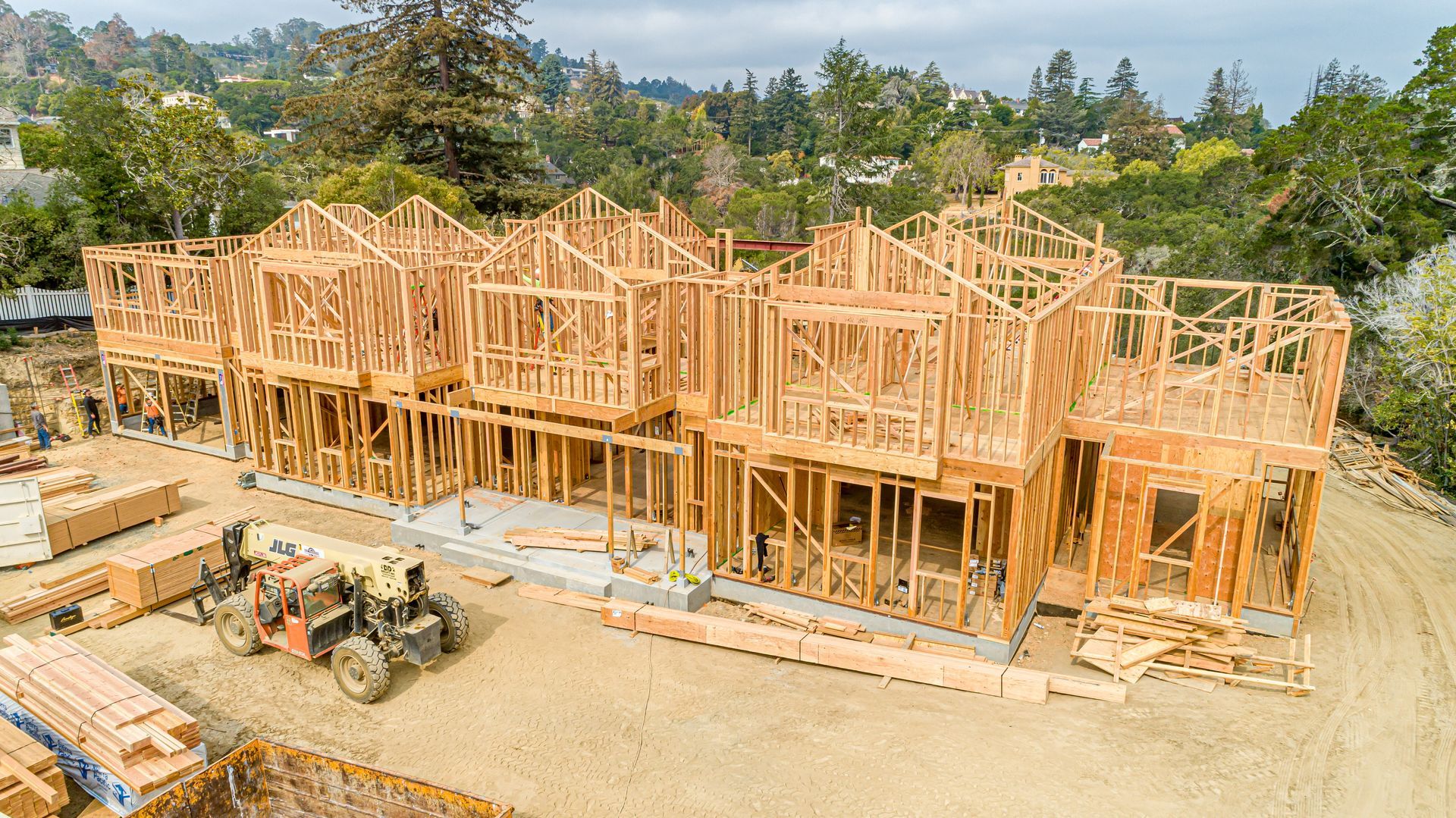 Framed wood construction of a large house on a dirt lot, with equipment and trees in the background