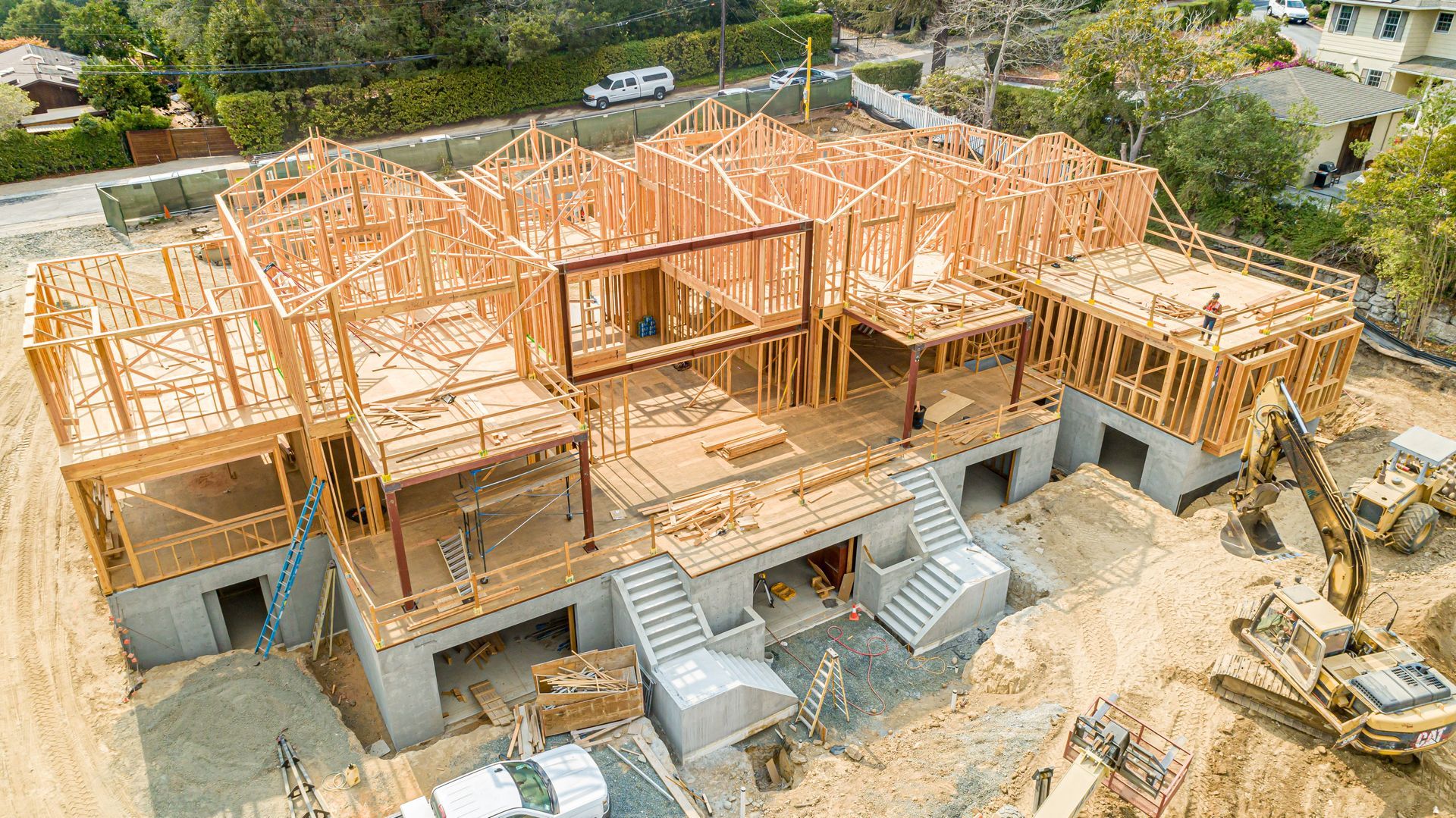 Aerial view of a house under construction with wooden framing, concrete walls, and heavy machinery nearby