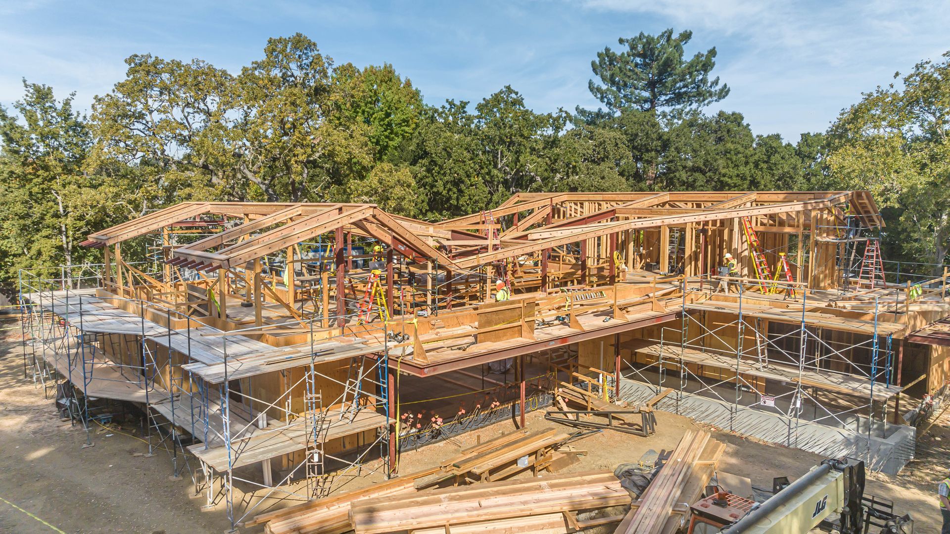 Wooden house frame under construction, surrounded by scaffolding and trees under a clear sky.
