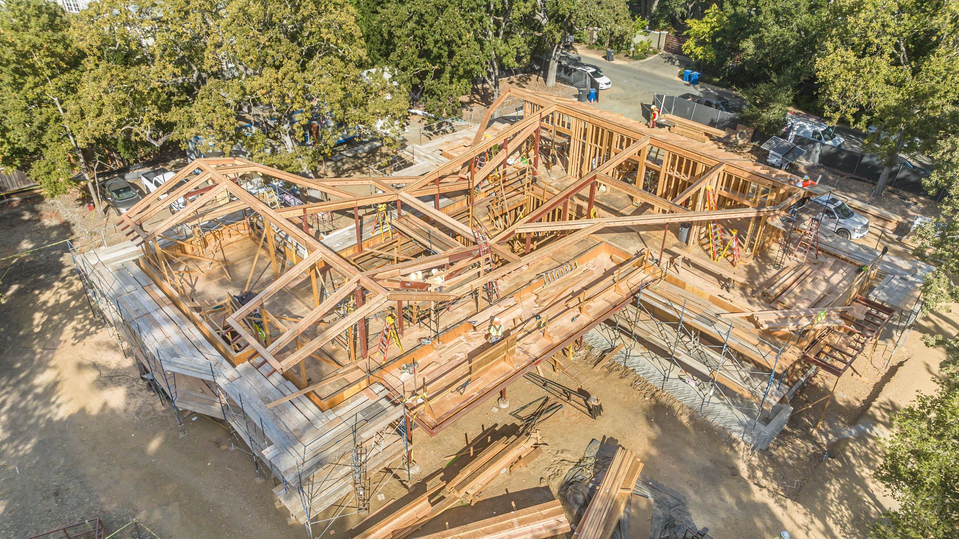 Aerial view of a wooden house frame under construction beside trees and dirt lot.
