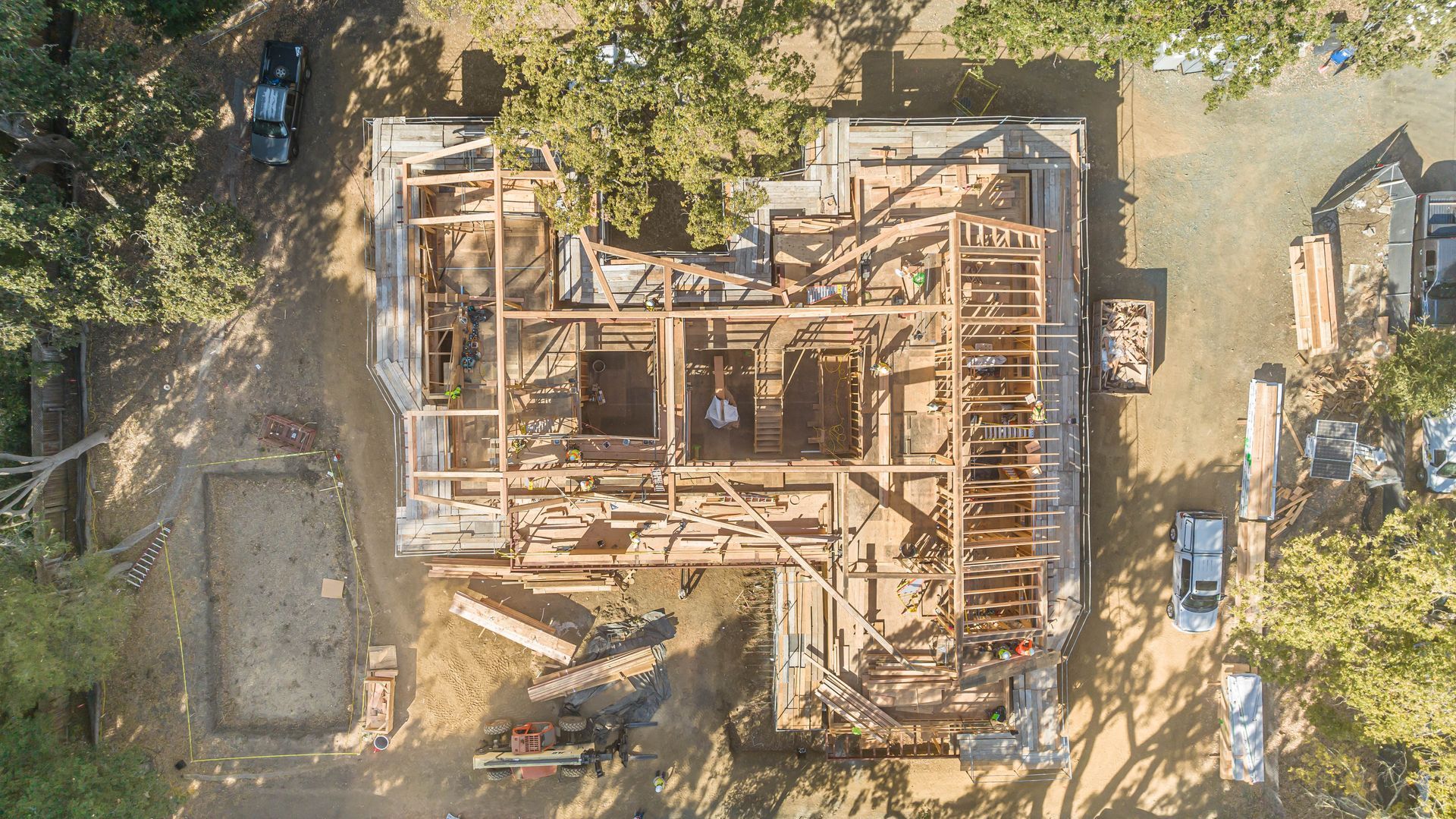 Aerial view of a house under construction with wooden framing, surrounded by dirt, trees, and construction materials