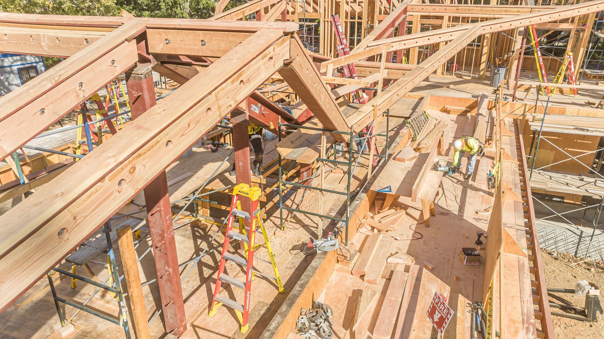 Wooden house framing under construction with workers, beams, and scaffolding in bright sunlight