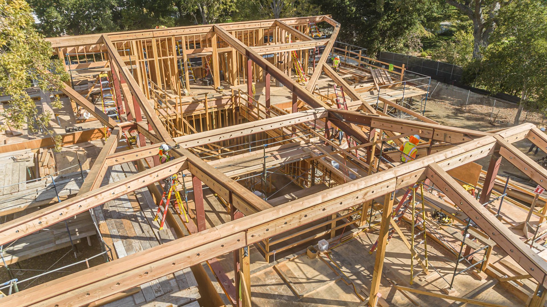Wooden house framing under construction on a sunny site, with beams, trusses, and exposed roof structure