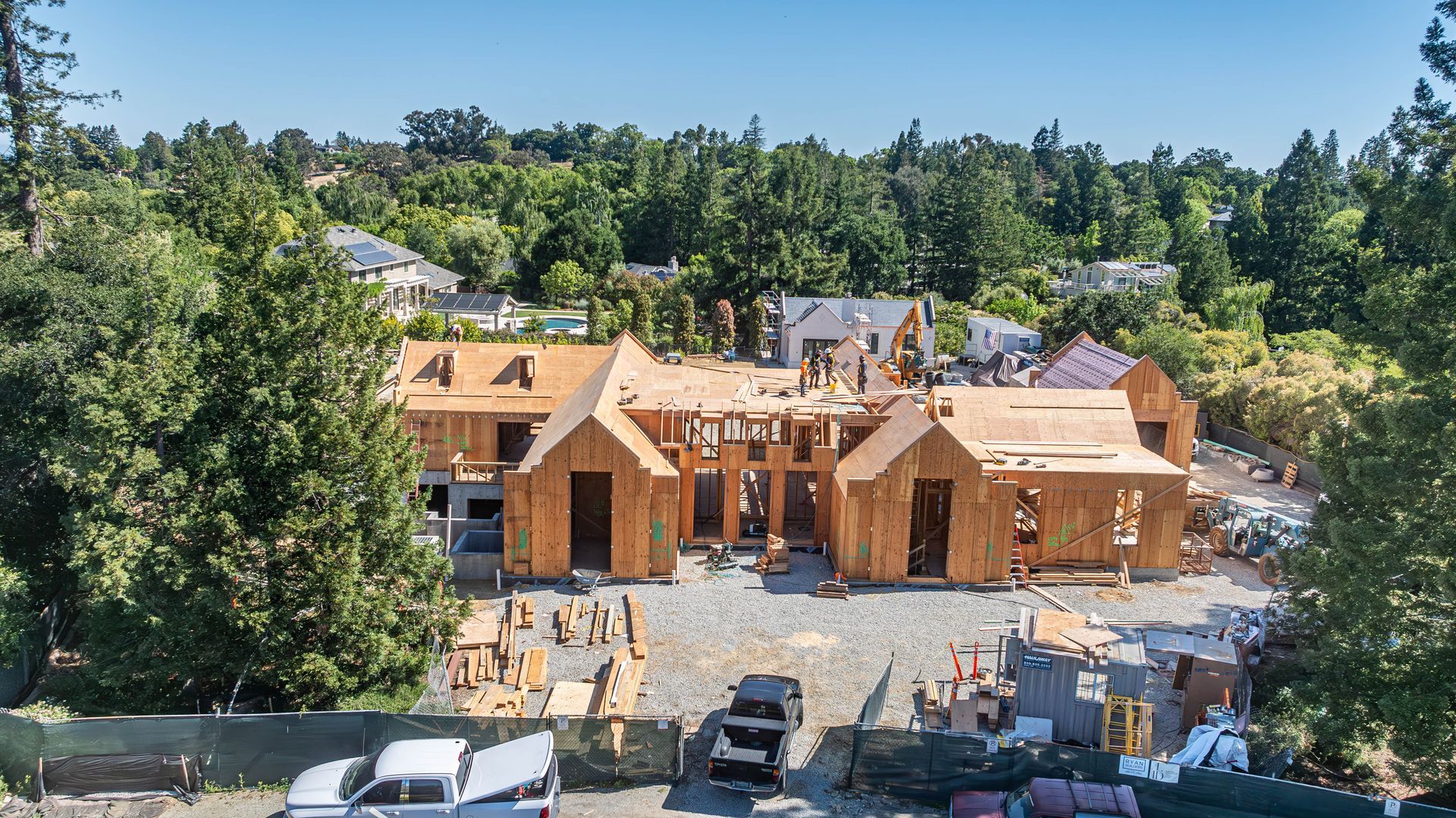 Wood-framed house under construction on a wooded hillside with parked vehicles in front