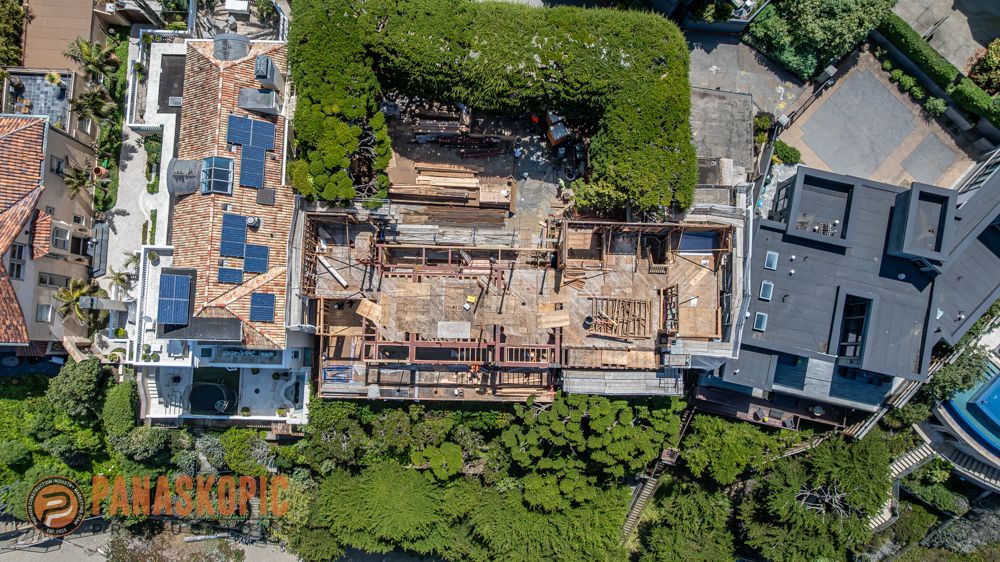 Aerial view of a building under construction beside a completed house and dense green trees.