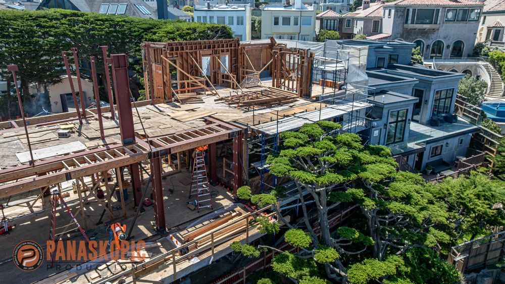 Construction site with wooden framing and scaffolding beside dense green trees and nearby buildings