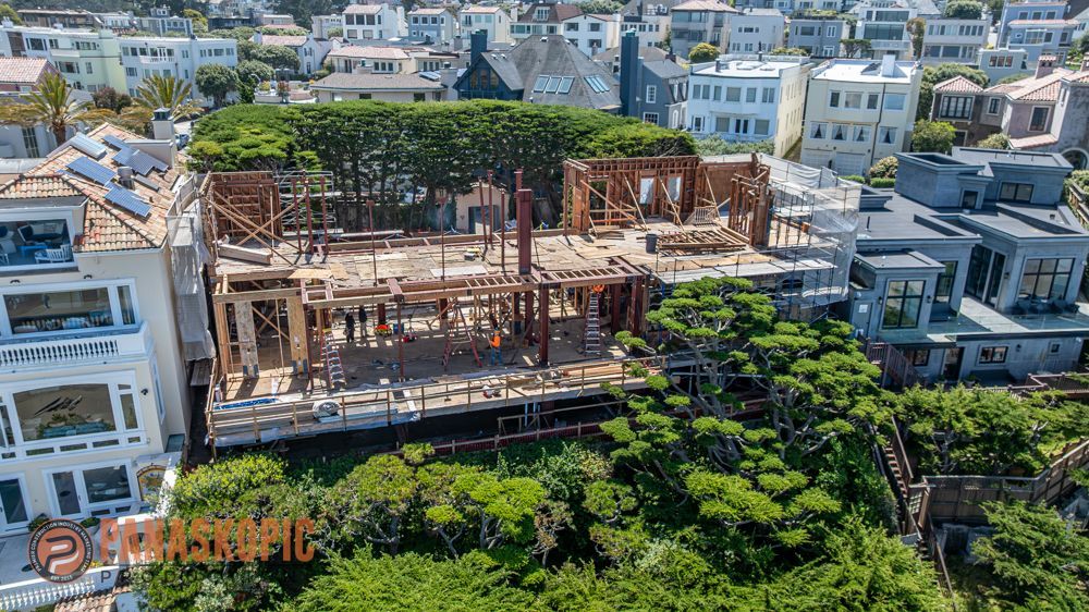 A wooden structure under construction beside a large green tree in a dense neighborhood of white buildings.