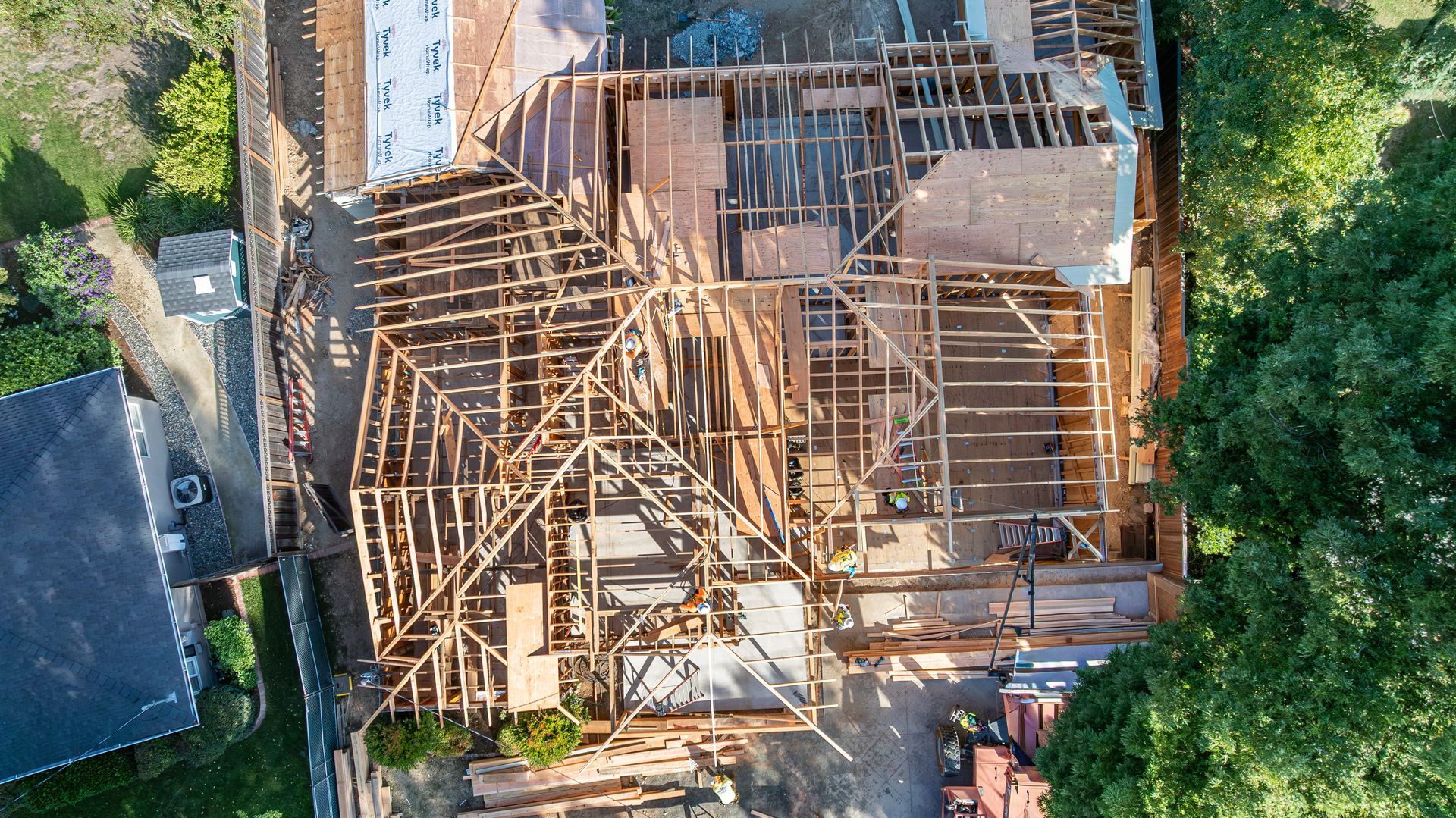 Aerial view of a house under construction with exposed wooden roof framing and surrounding trees.