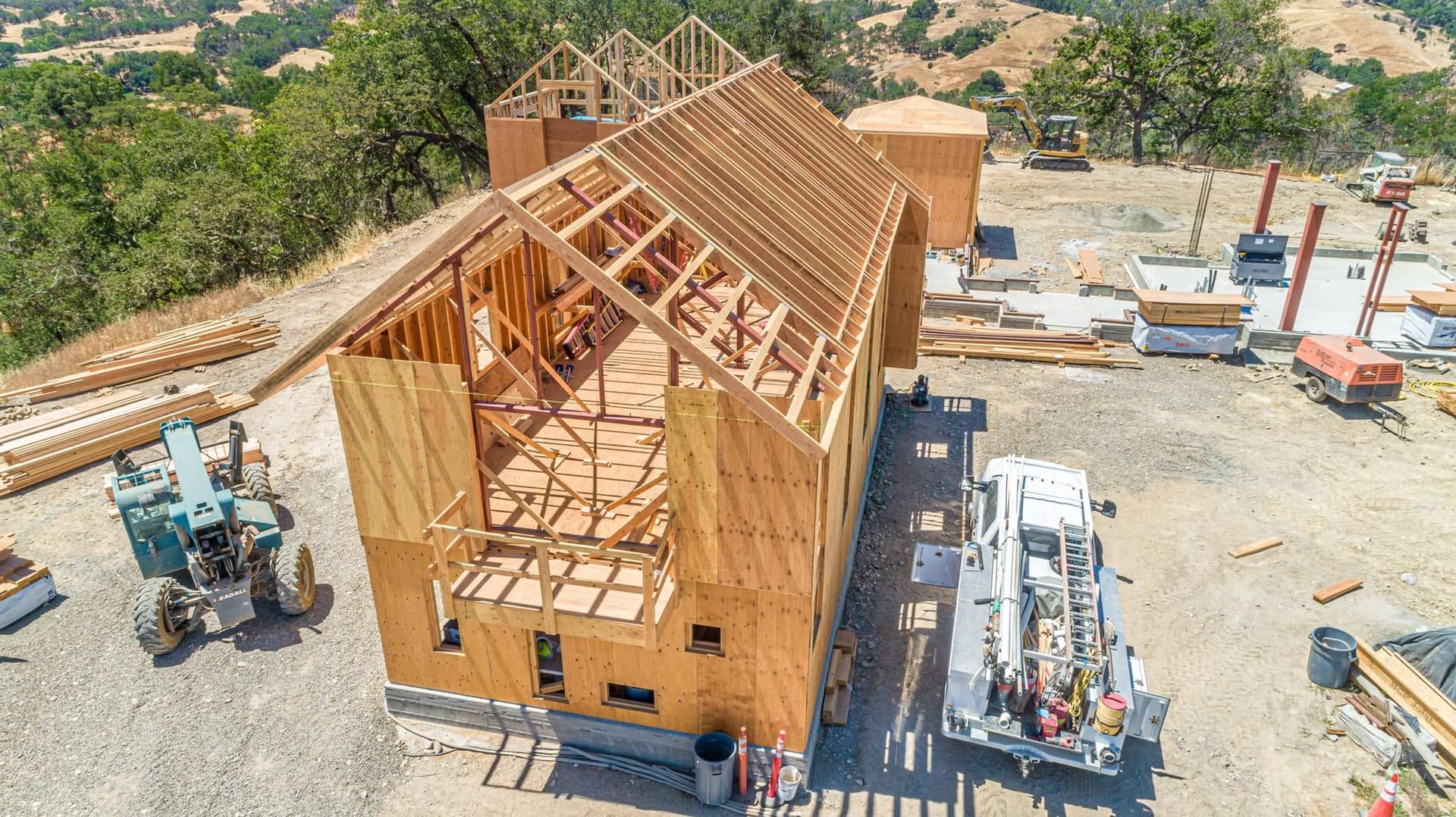 Aerial view of a wooden house under construction with roof framing and construction vehicles on a dirt lot.