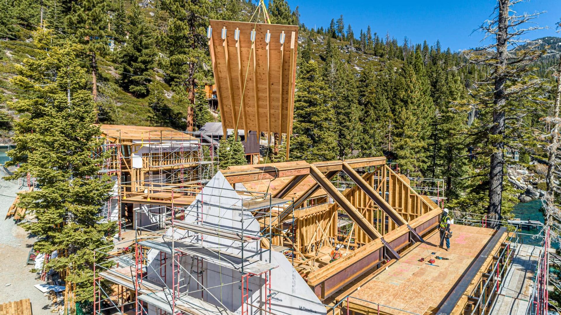 Wooden house under construction in a forest, with exposed roof framing and a tall chimney stack.