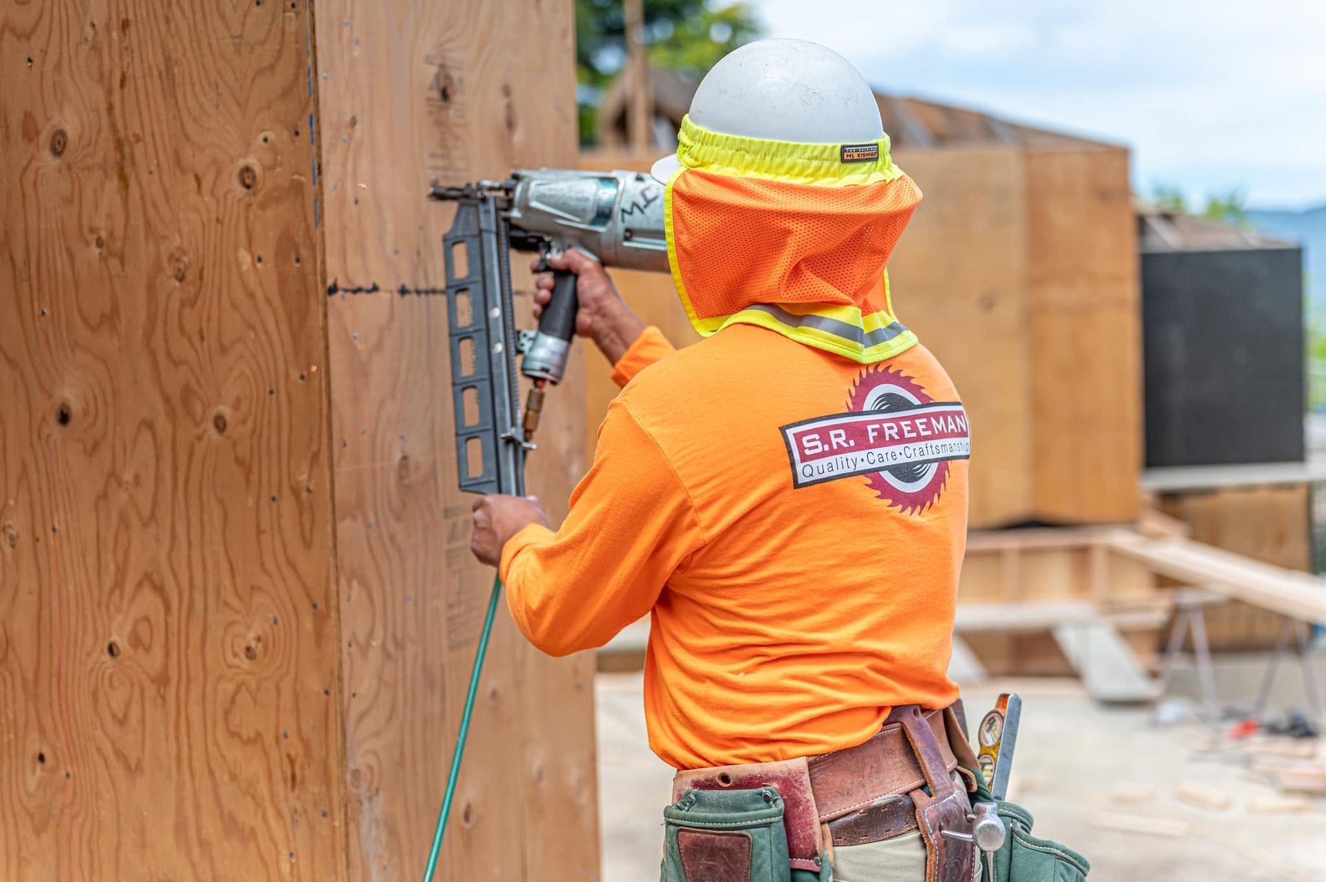 Worker in orange shirt fastening plywood to an outdoor wall with a nail gun.