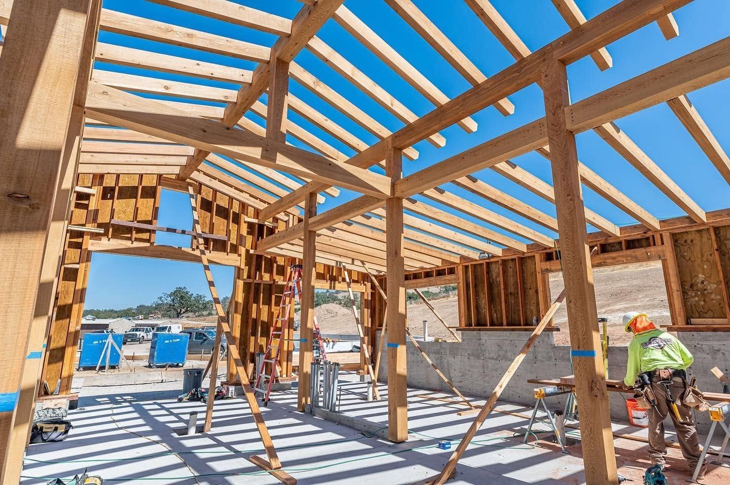 Wood-framed house under construction, with workers, blue barrels, and bright sunlight through roof beams