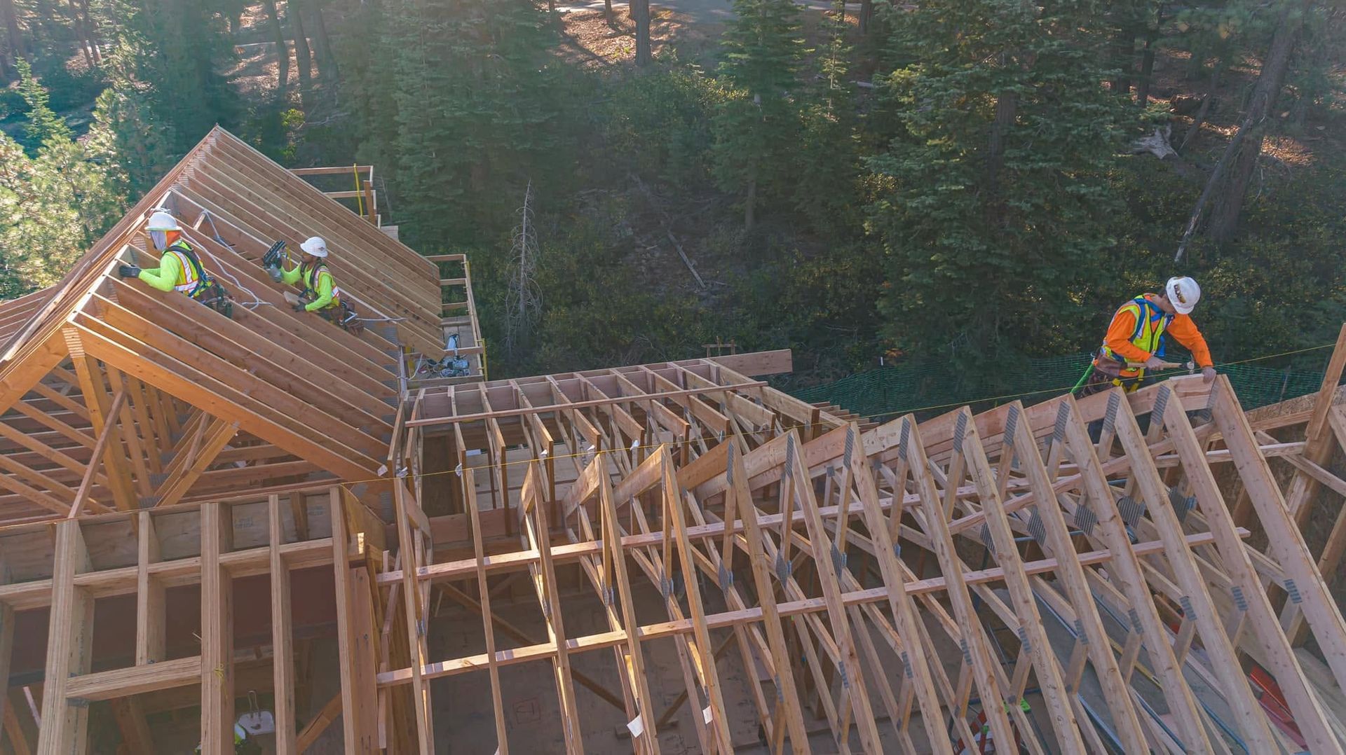 Workers building a wooden roof frame in a forest clearing, with one section partly assembled.