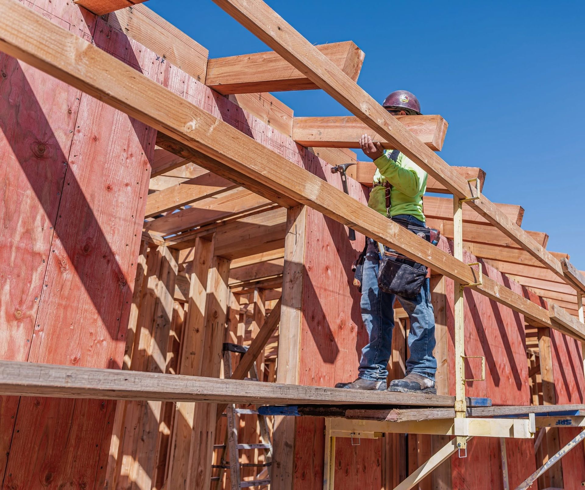 Worker framing a house on a wooden scaffold at a construction site under a clear blue sky