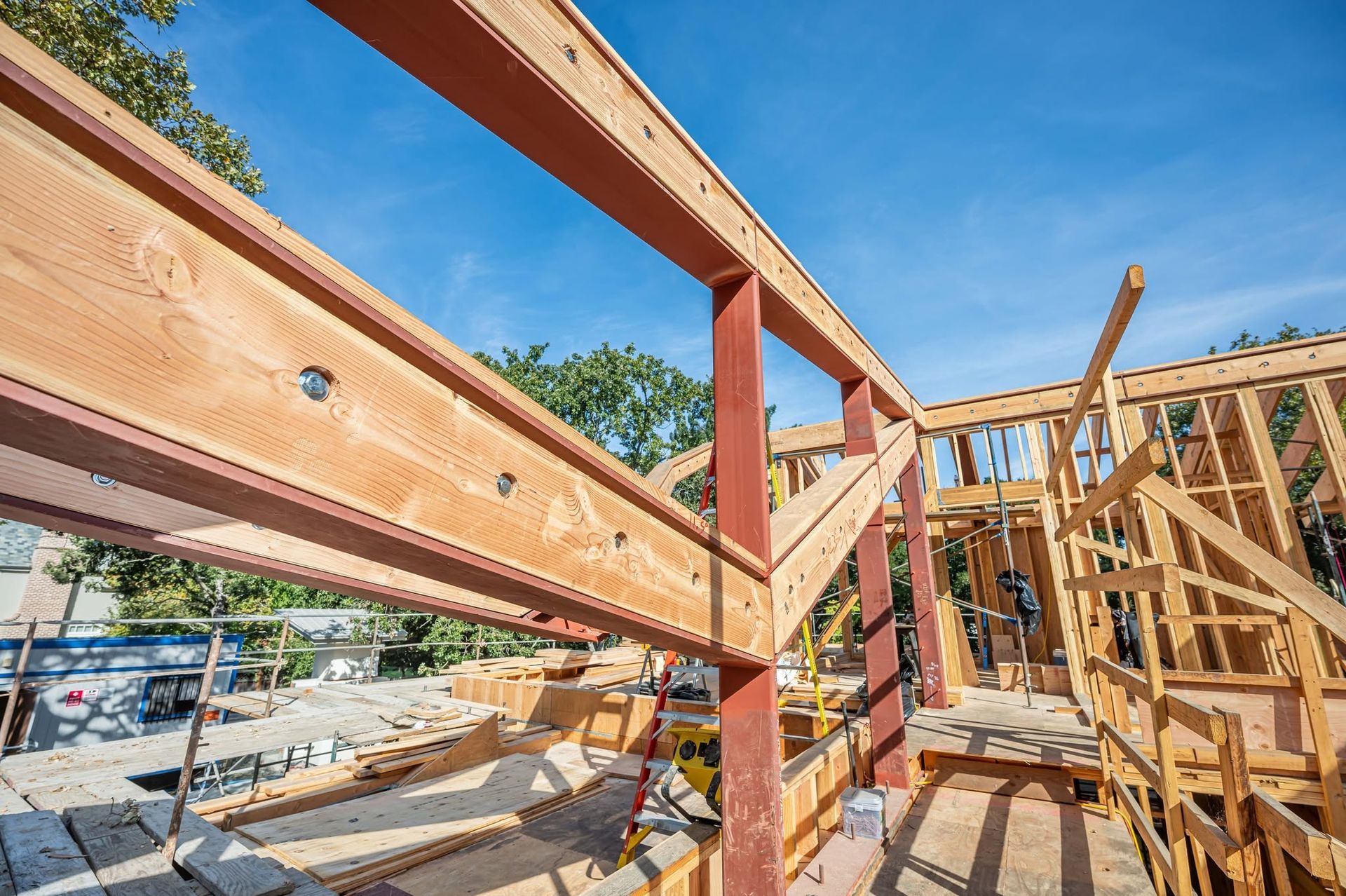 Wooden house framing under construction against a blue sky, with beams and scaffolding visible.